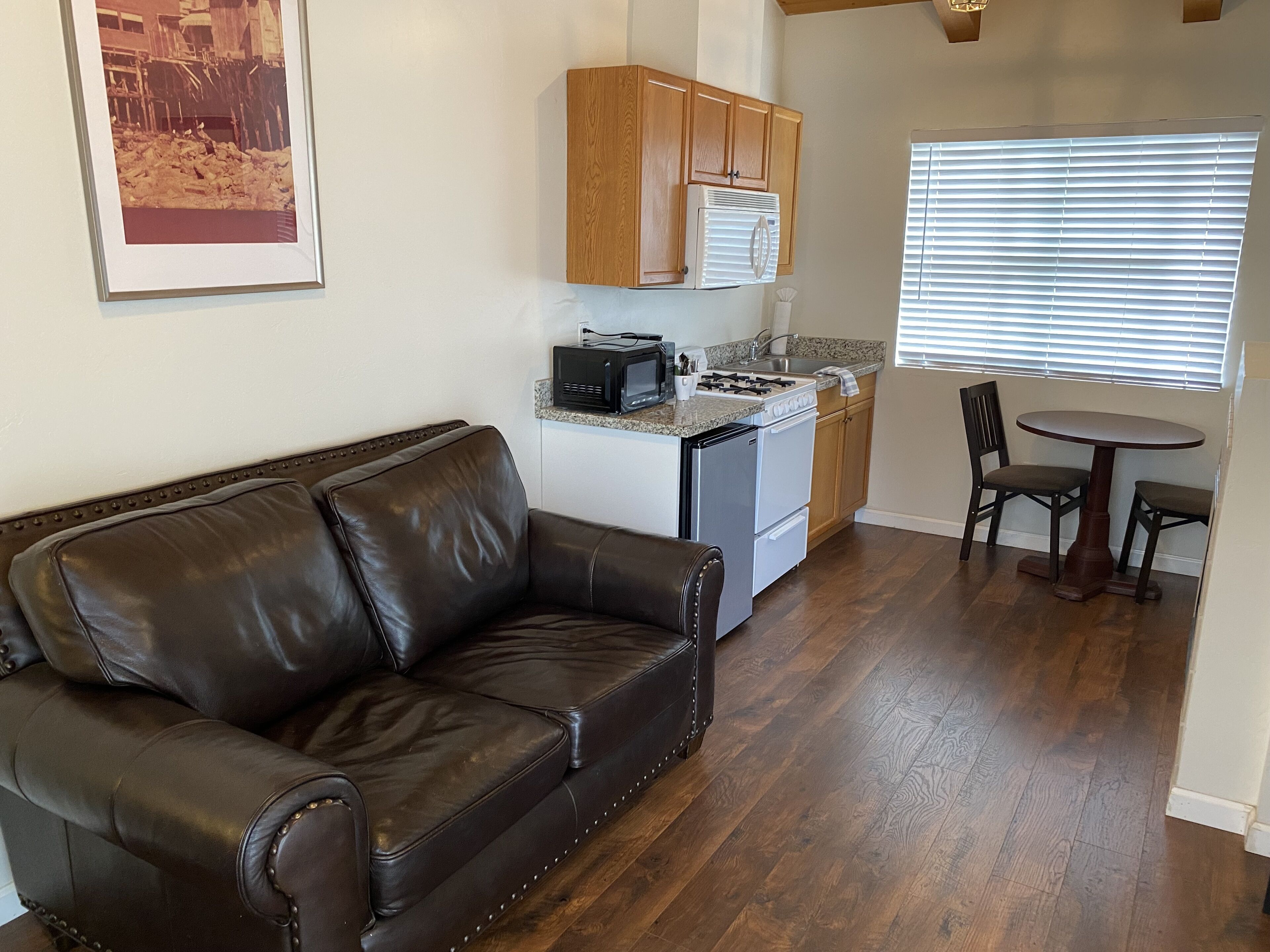 A compact living space with a brown leather couch, a kitchenette with a stove, microwave, and refrigerator, and a dining area with two chairs and a table.