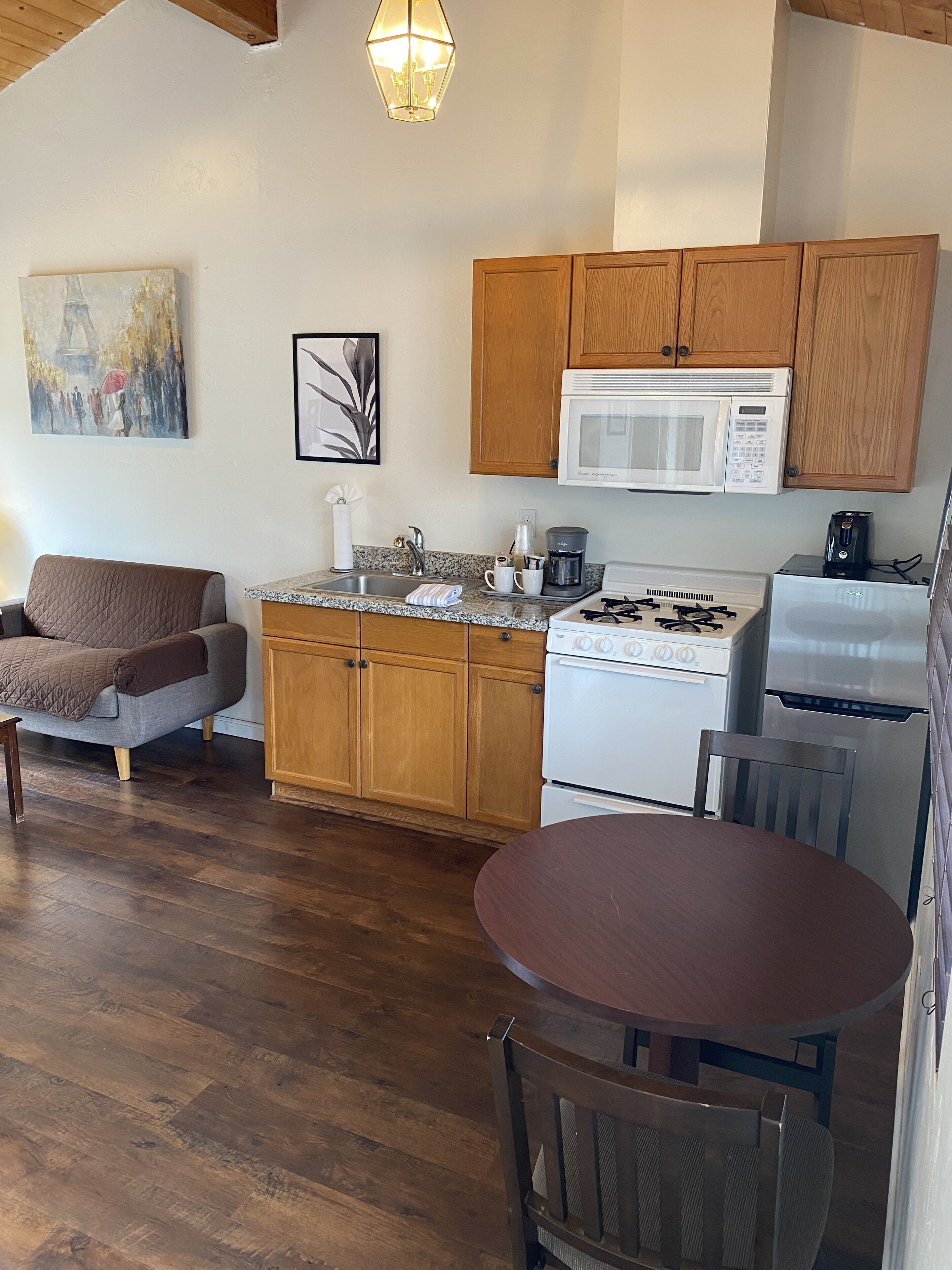 A cozy kitchen with wooden cabinets, a white stove, a microwave, and a coffee maker. There is a dining table with chairs and a couch in the adjacent living area.