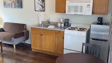 A cozy kitchen with wooden cabinets, a white stove, a microwave, and a coffee maker. There is a dining table with chairs and a couch in the adjacent living area.