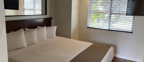 A bedroom with a large bed, a headboard, a window with blinds, and a mirror on the wall.