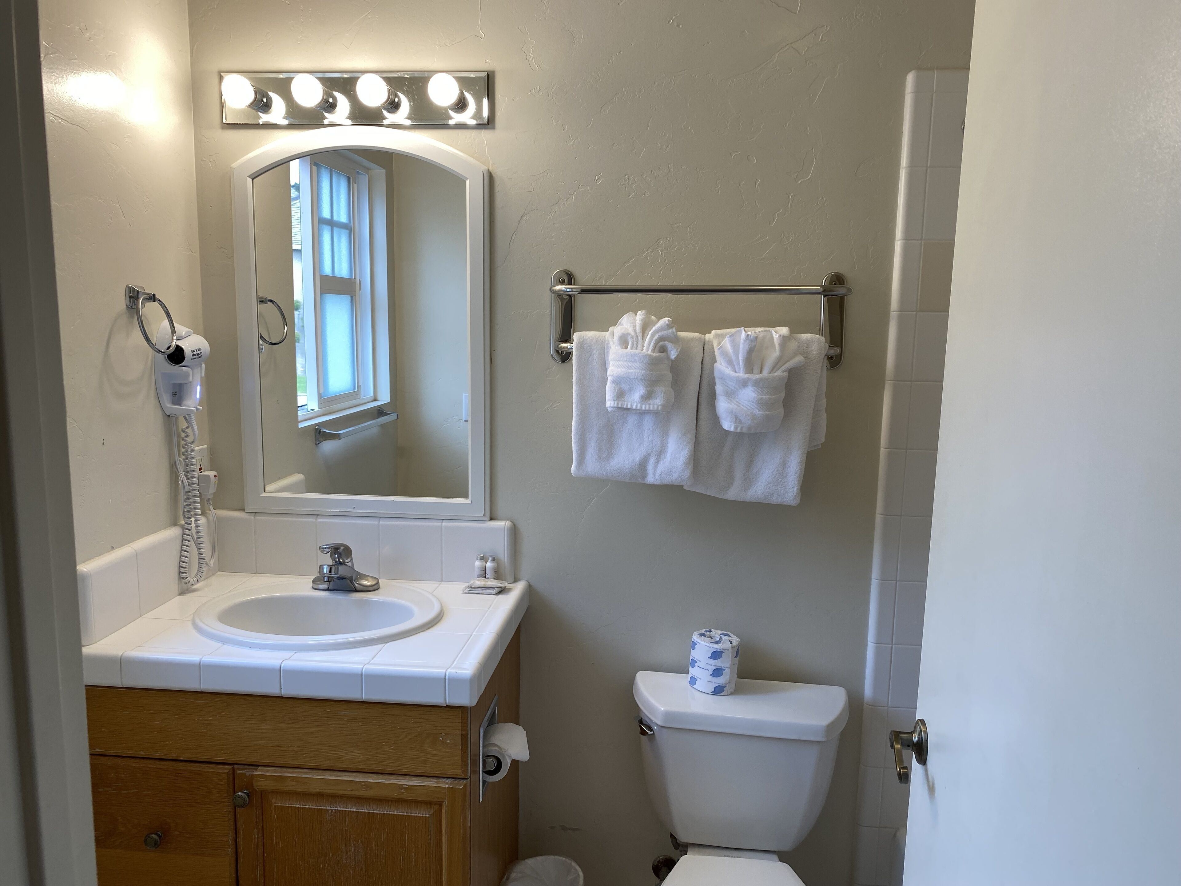 Une salle de bain équipée d’un lavabo, d’un miroir, d’un sèche-cheveux, d’un porte-serviettes, de toilettes et d’un rouleau de papier toilette.