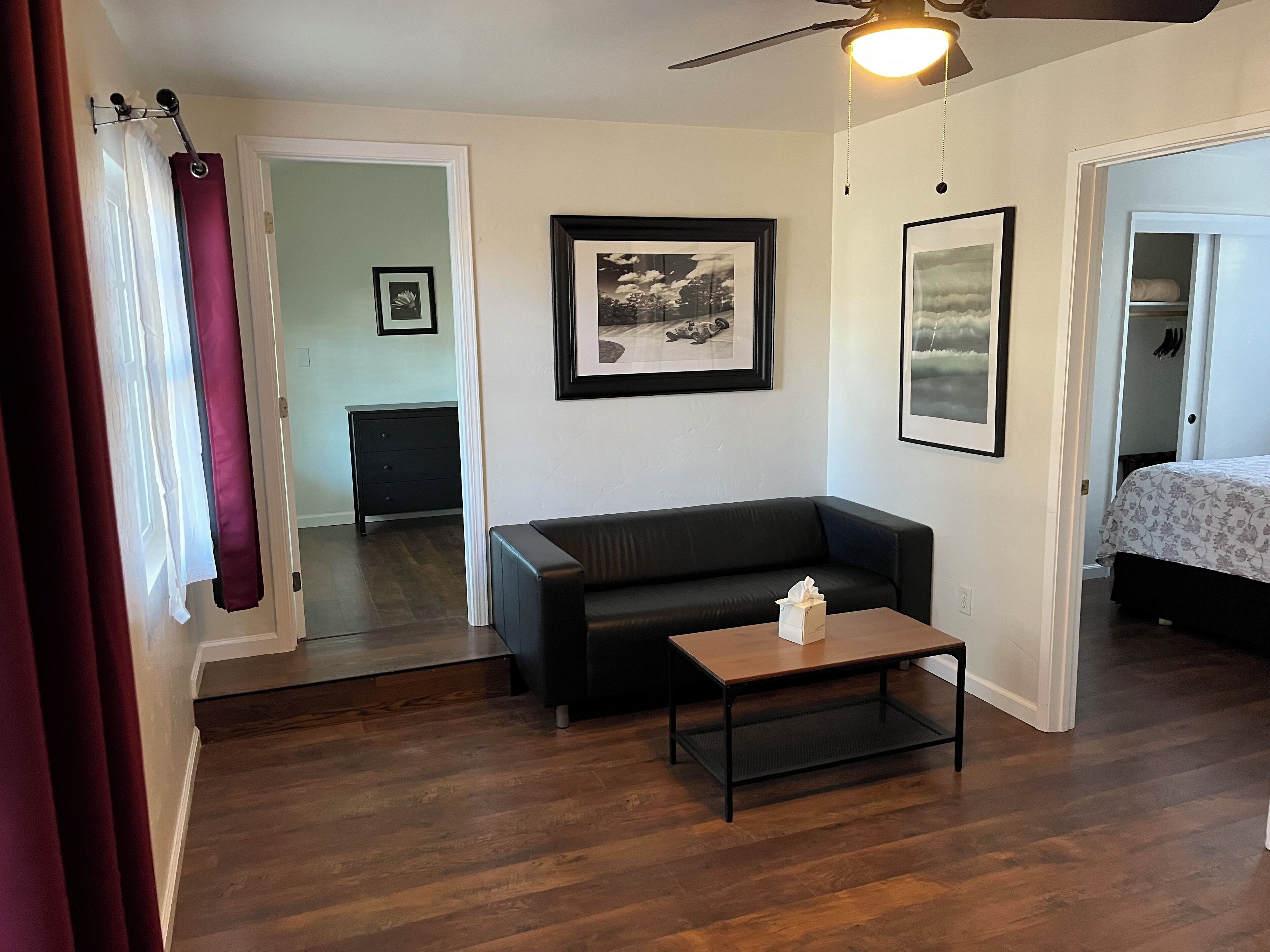 Una habitación con un sofá de cuero negro, una mesa de centro de madera y un ventilador de techo. Hay cuadros enmarcados en las paredes y una puerta que da a otra habitación.