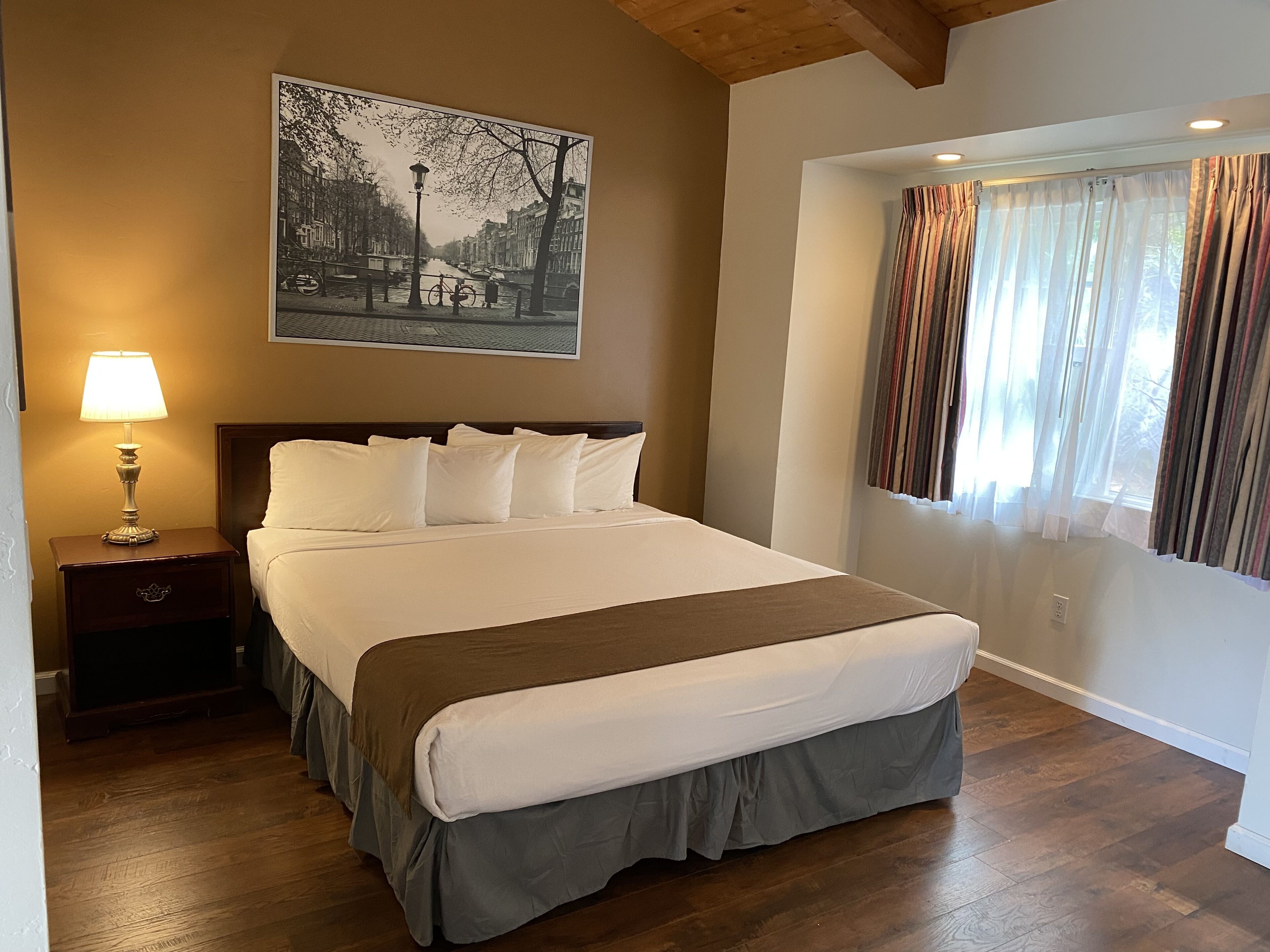 A neatly made bed with white linens and a brown accent, a bedside lamp, and a framed black and white cityscape painting on the wall.