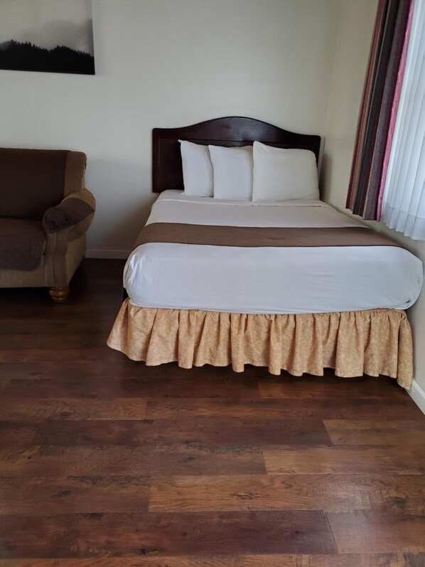 A neatly made bed with a wooden headboard, a brown skirted bedspread, and a brown chair to the left.