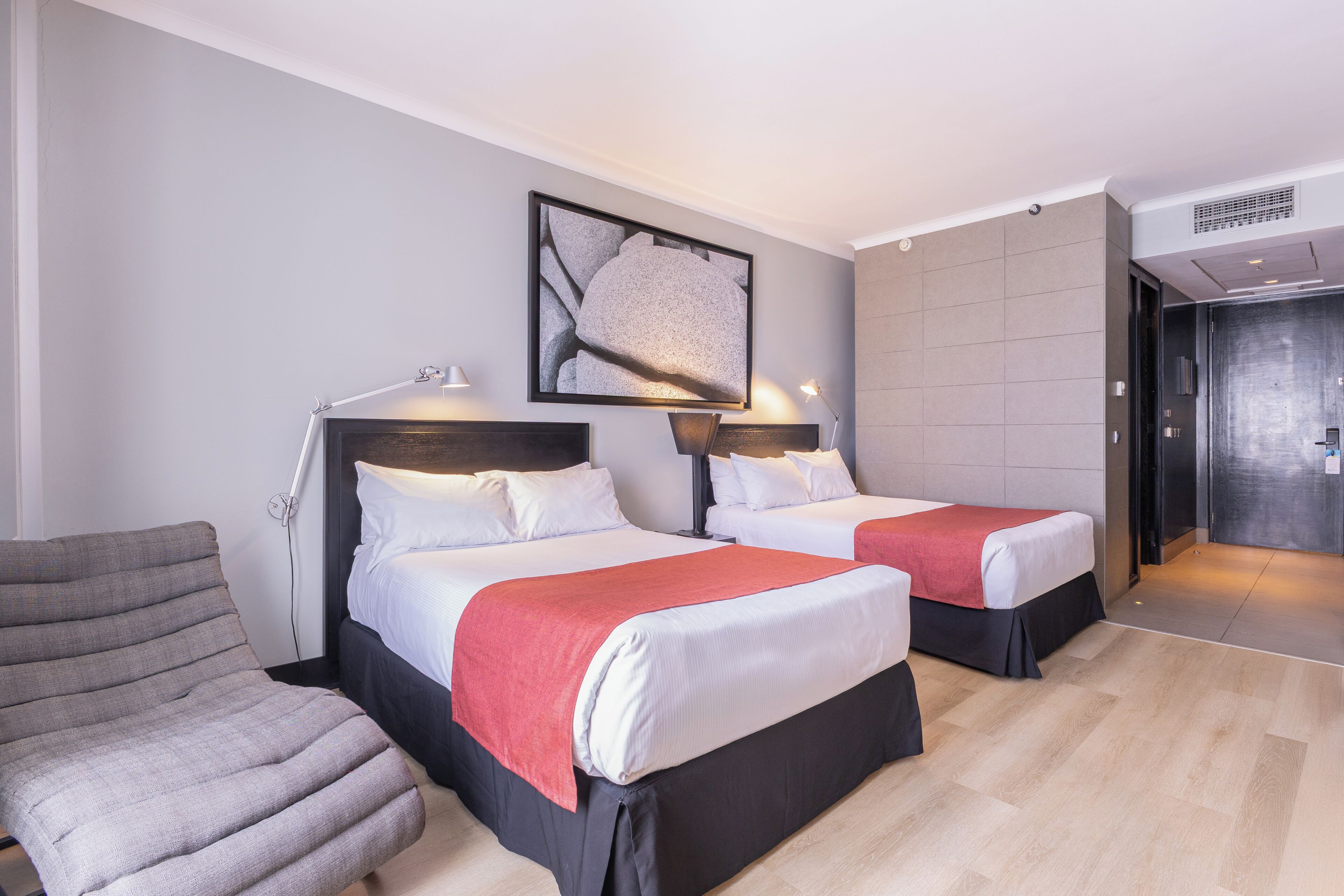 Habitación de hotel con dos camas, una silla, una obra de arte enmarcada y una puerta que da a un baño.