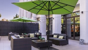 Un área de estar al aire libre con mobiliario de mimbre, sombrillas verdes y un edificio moderno al fondo.