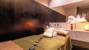 A spa room with a massage table, a wall-mounted massage head, a bottle of oil, and a towel.