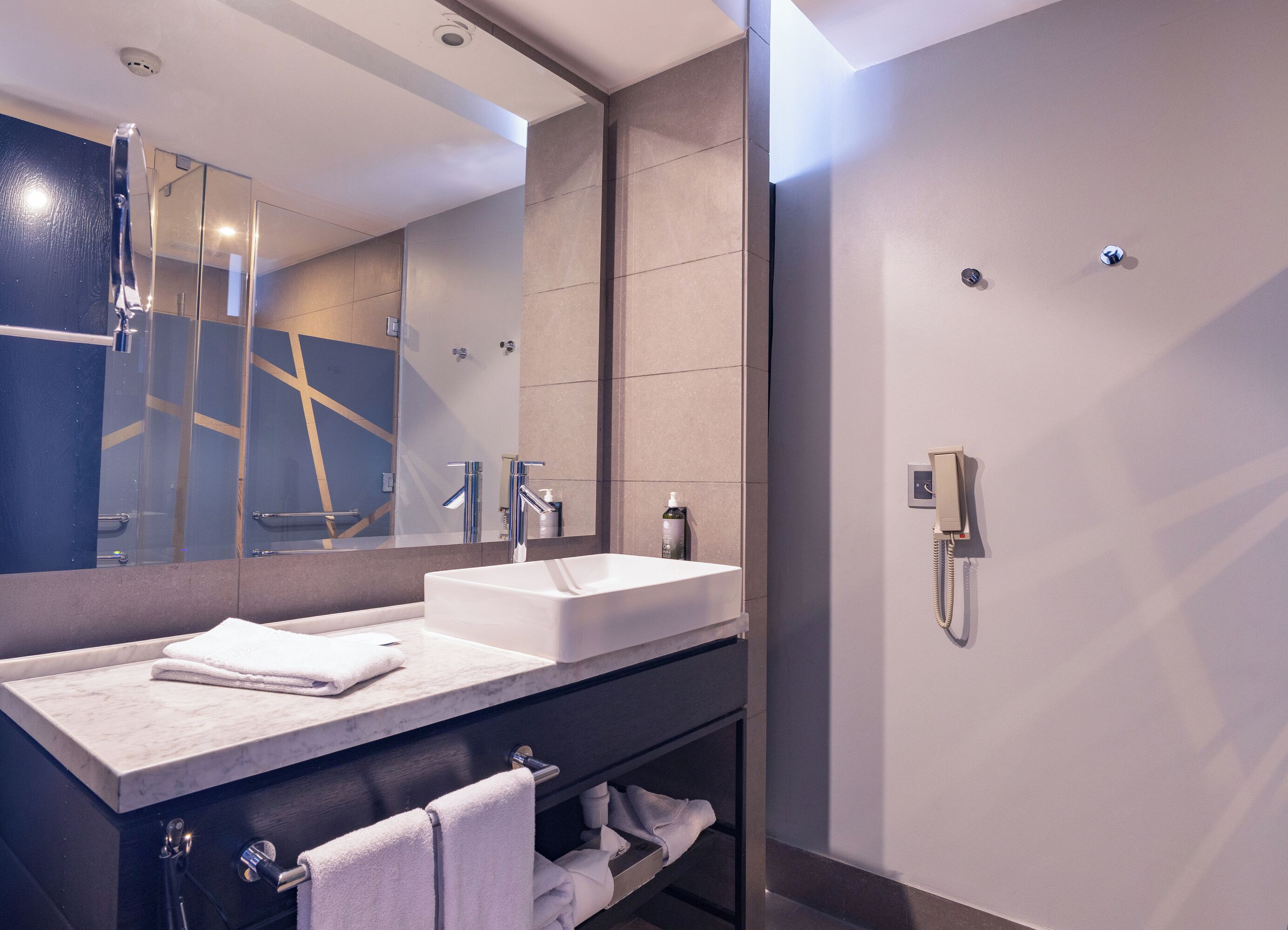 A modern bathroom with a large mirror, two sinks, a towel rack, and a shower.