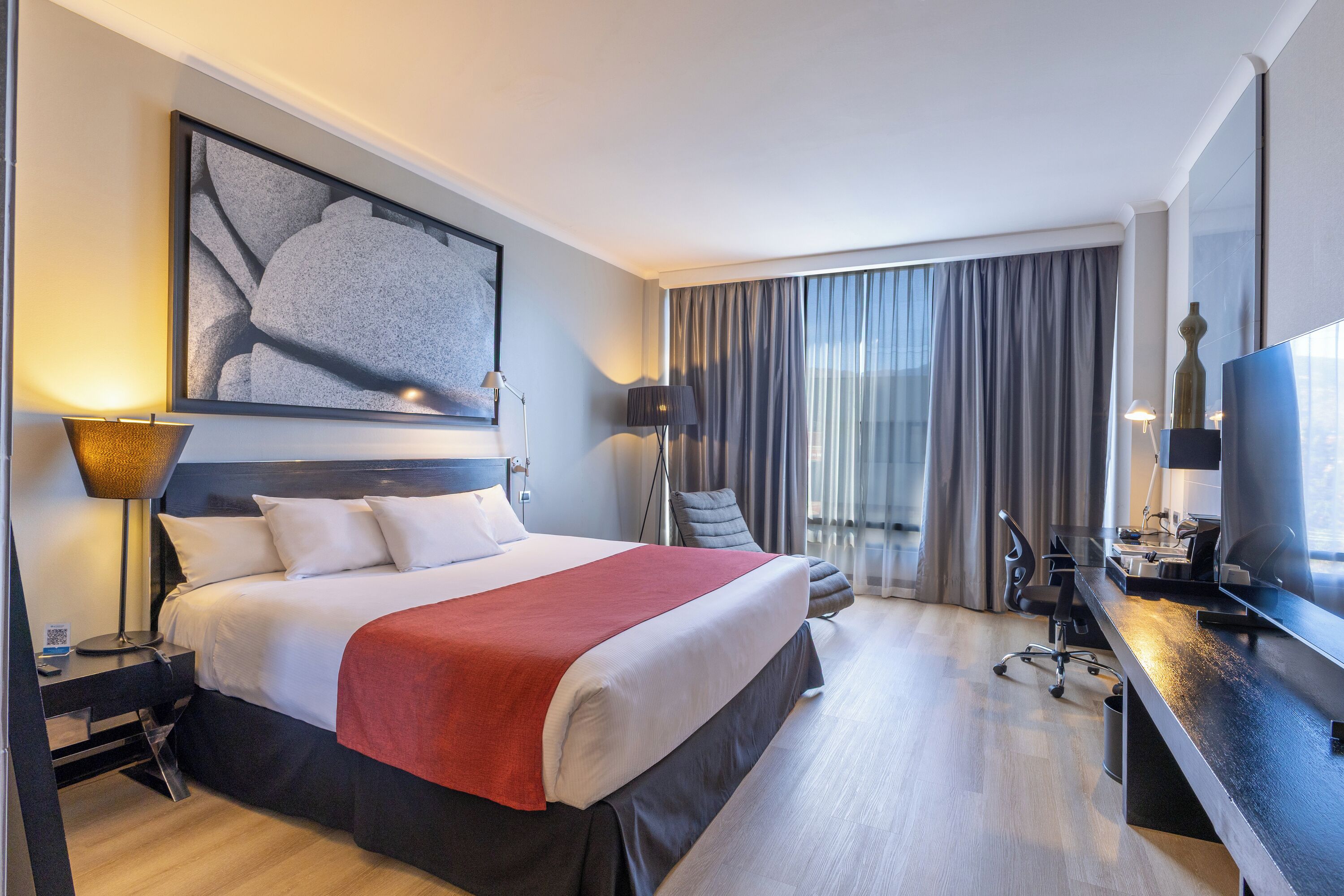 A hotel room with a large bed, a desk with a computer, a chair, a lamp, and a painting on the wall.
