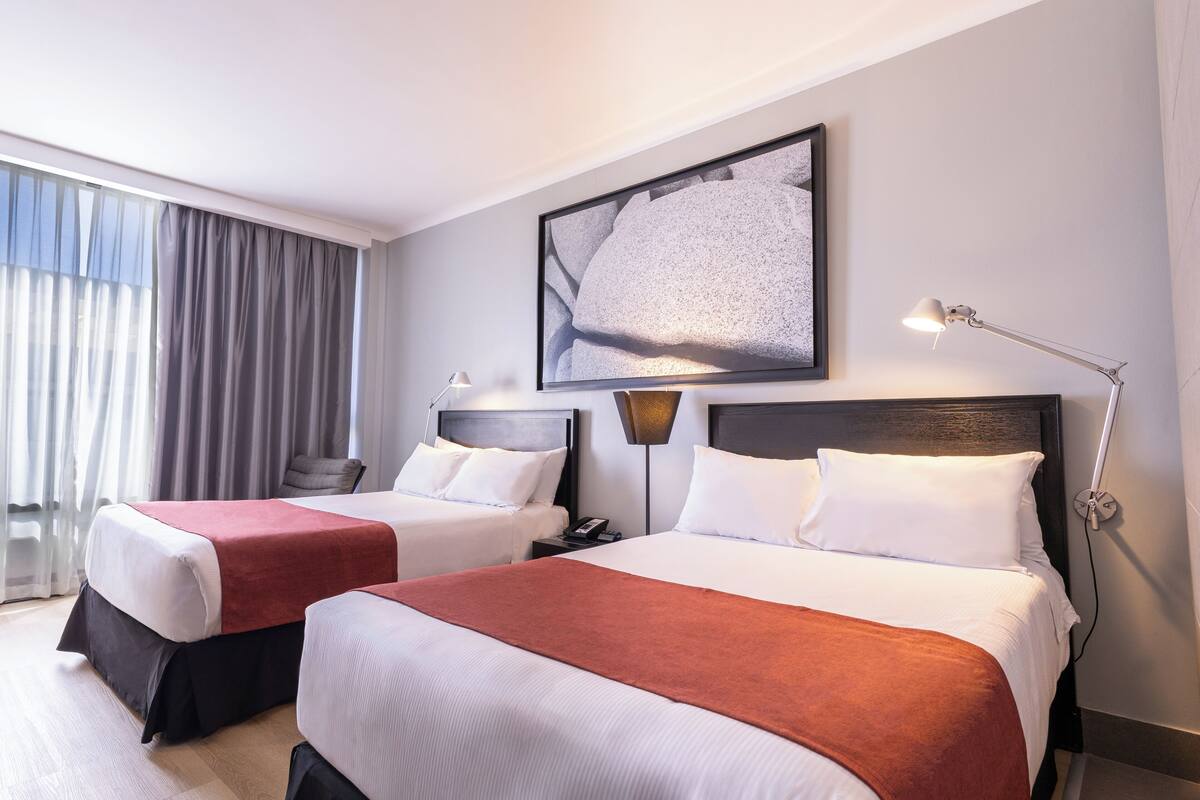 Een hotelkamer met twee bedden, een groot raam met gordijnen, een kunstwerk aan de muur en een bedlamp.