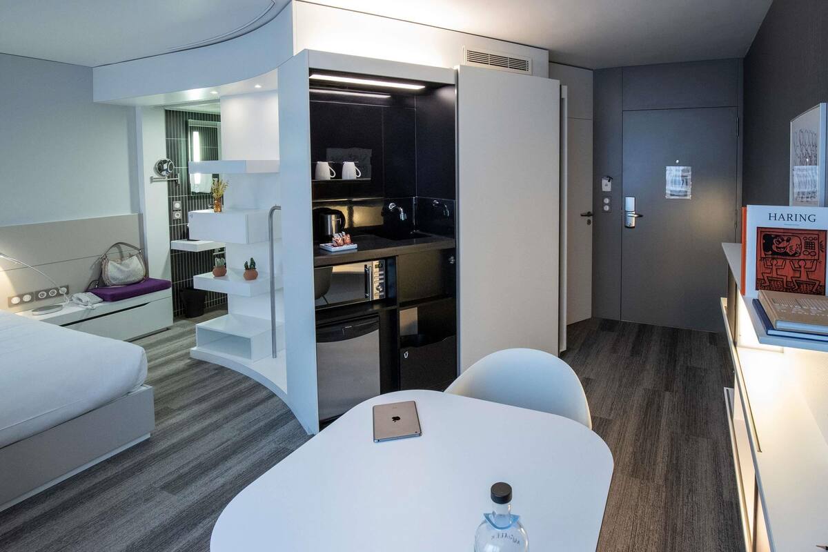 A modern hotel room with a kitchenette, a bed, a table with a bottle, and a chair.