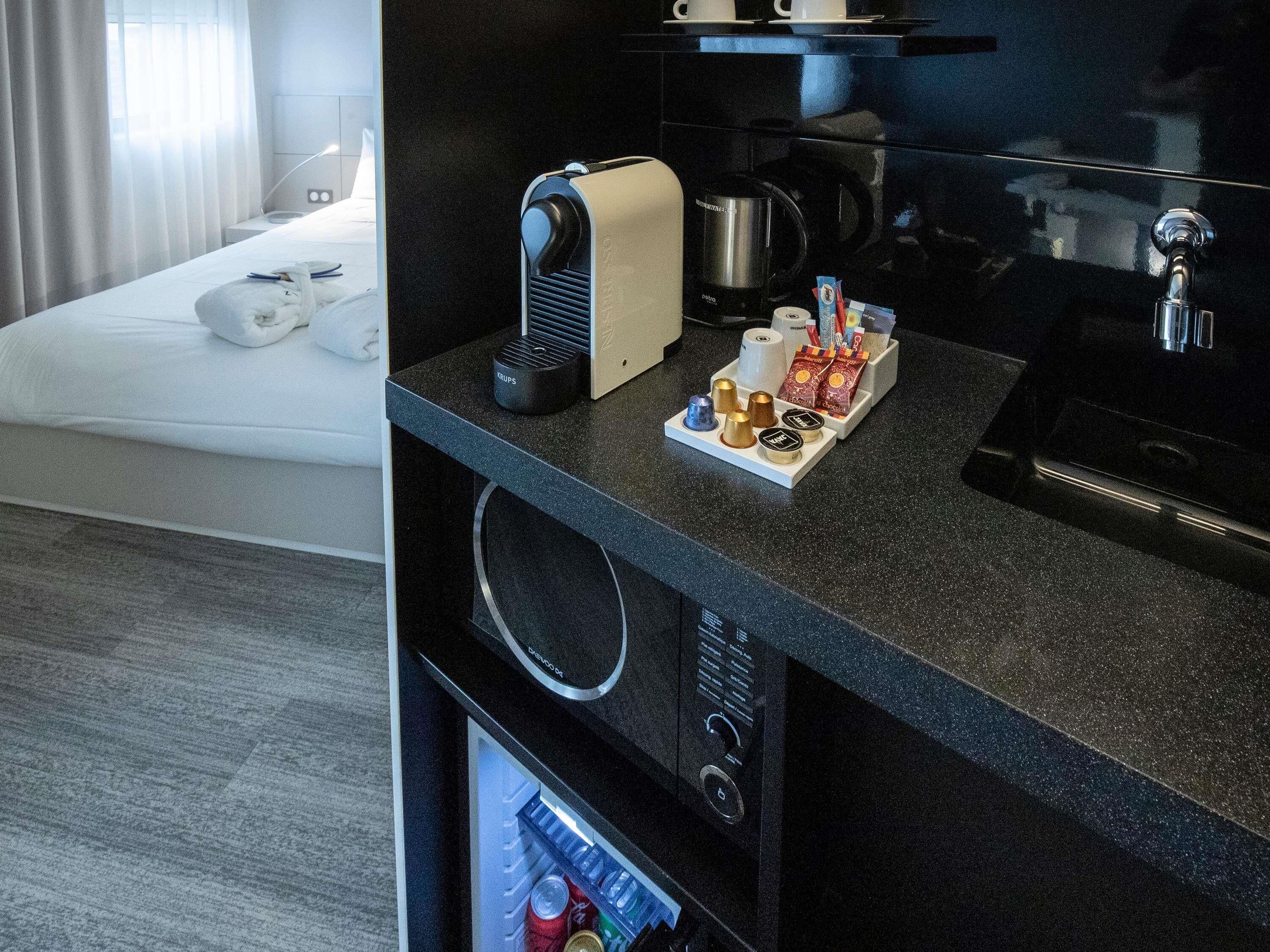 A modern hotel room with a bed, a minibar, a coffee maker, and a refrigerator.