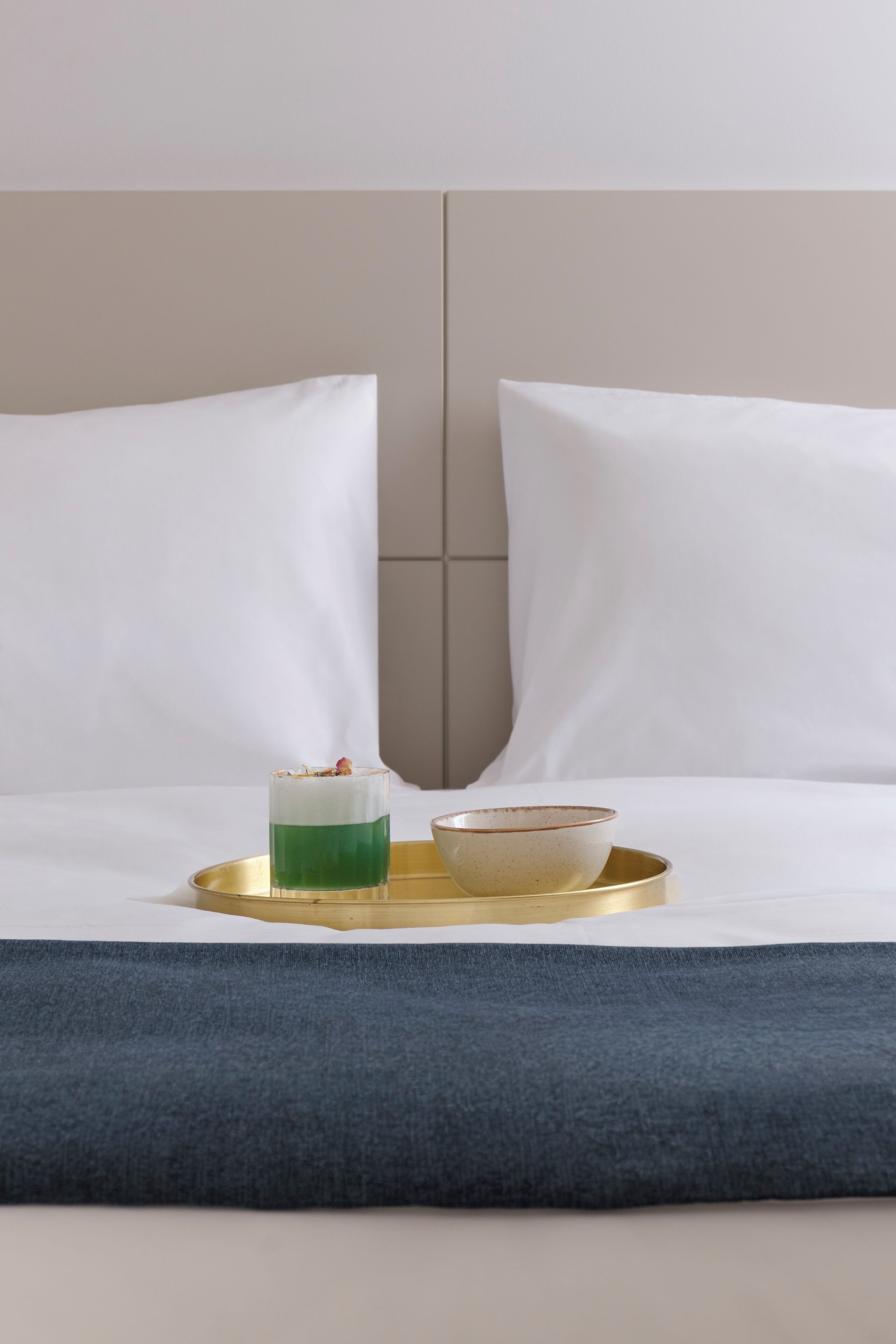 A bed with white pillows and a blue throw, a tray with a green drink and a bowl, and a wall with a vertical panel.