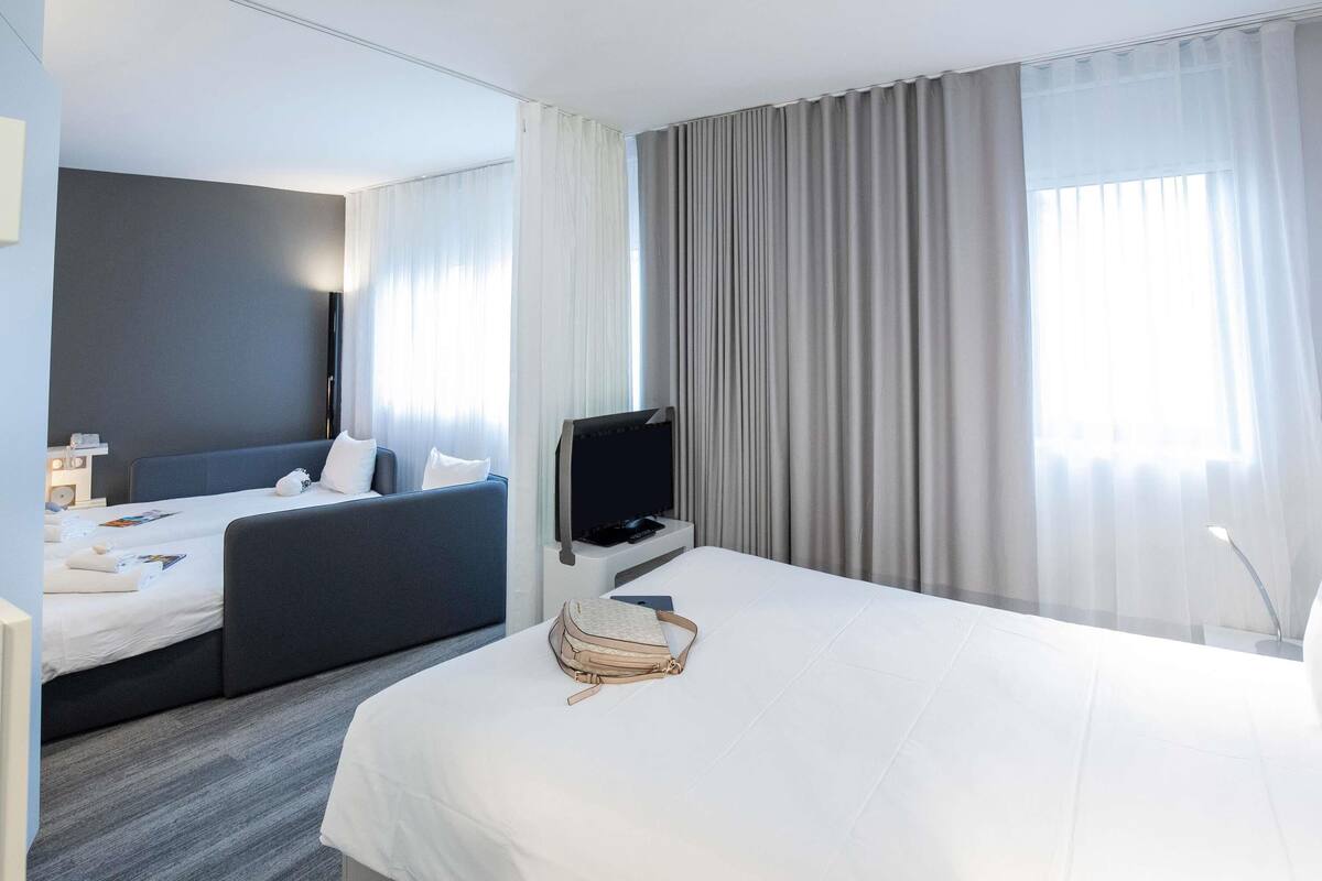 A hotel room with a large bed, a desk with a computer, a TV, and a seating area with a sofa and pillows.