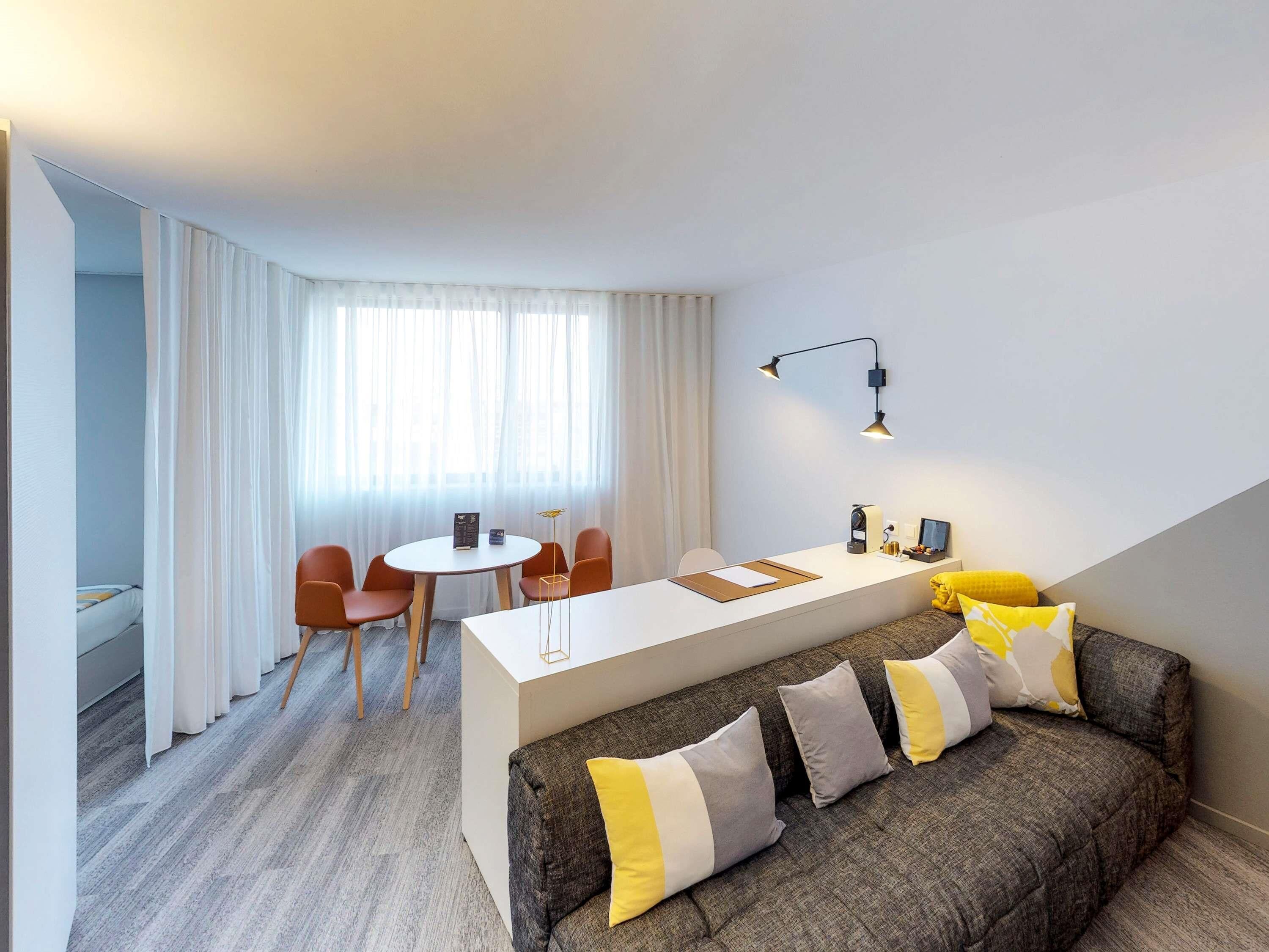 Una habitación de hotel moderna con zona de comedor, un sofá con cojines estampados y un escritorio con lámpara.