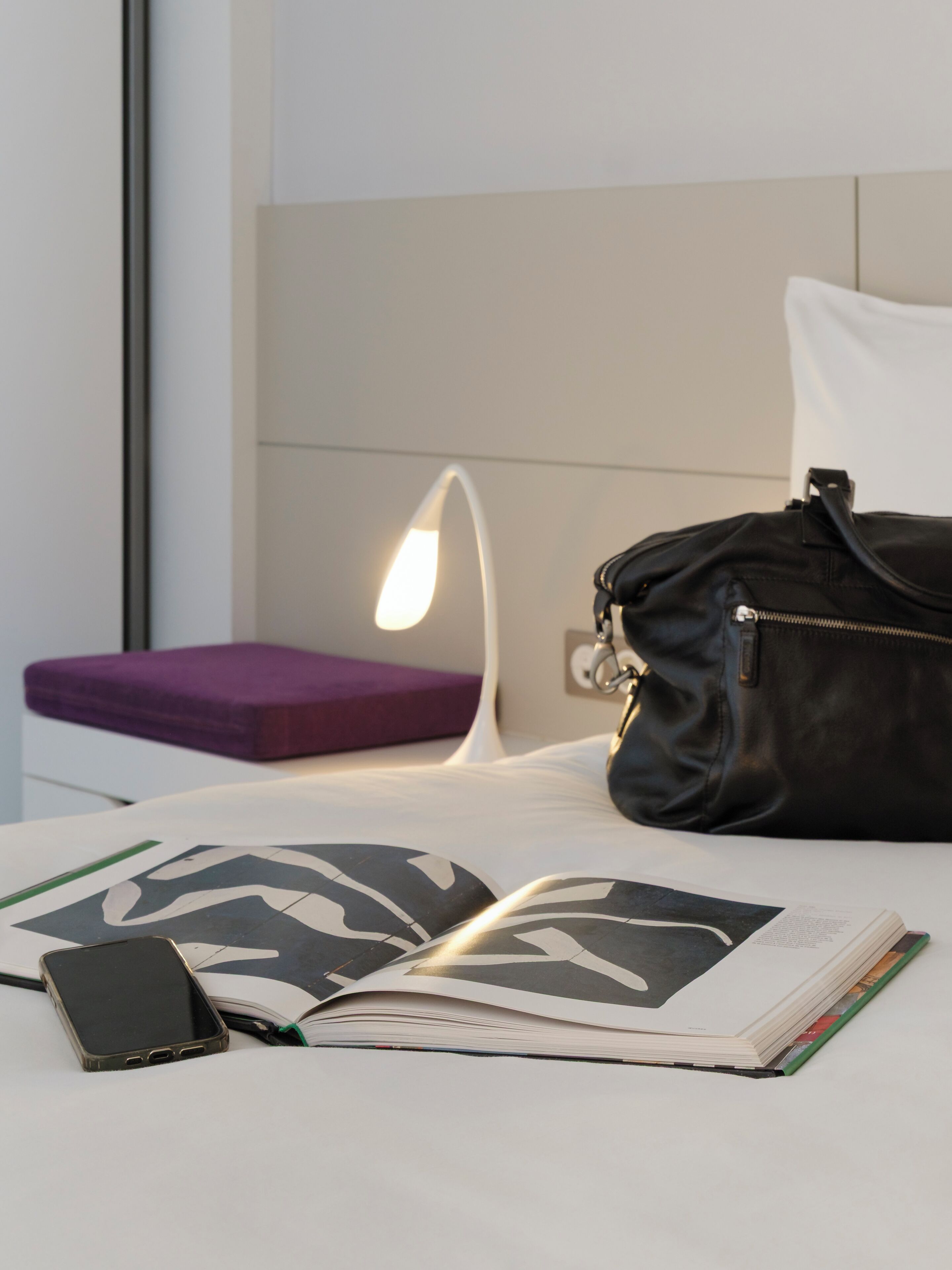 A bed with a black leather bag, a smartphone, and an open book on a white surface.