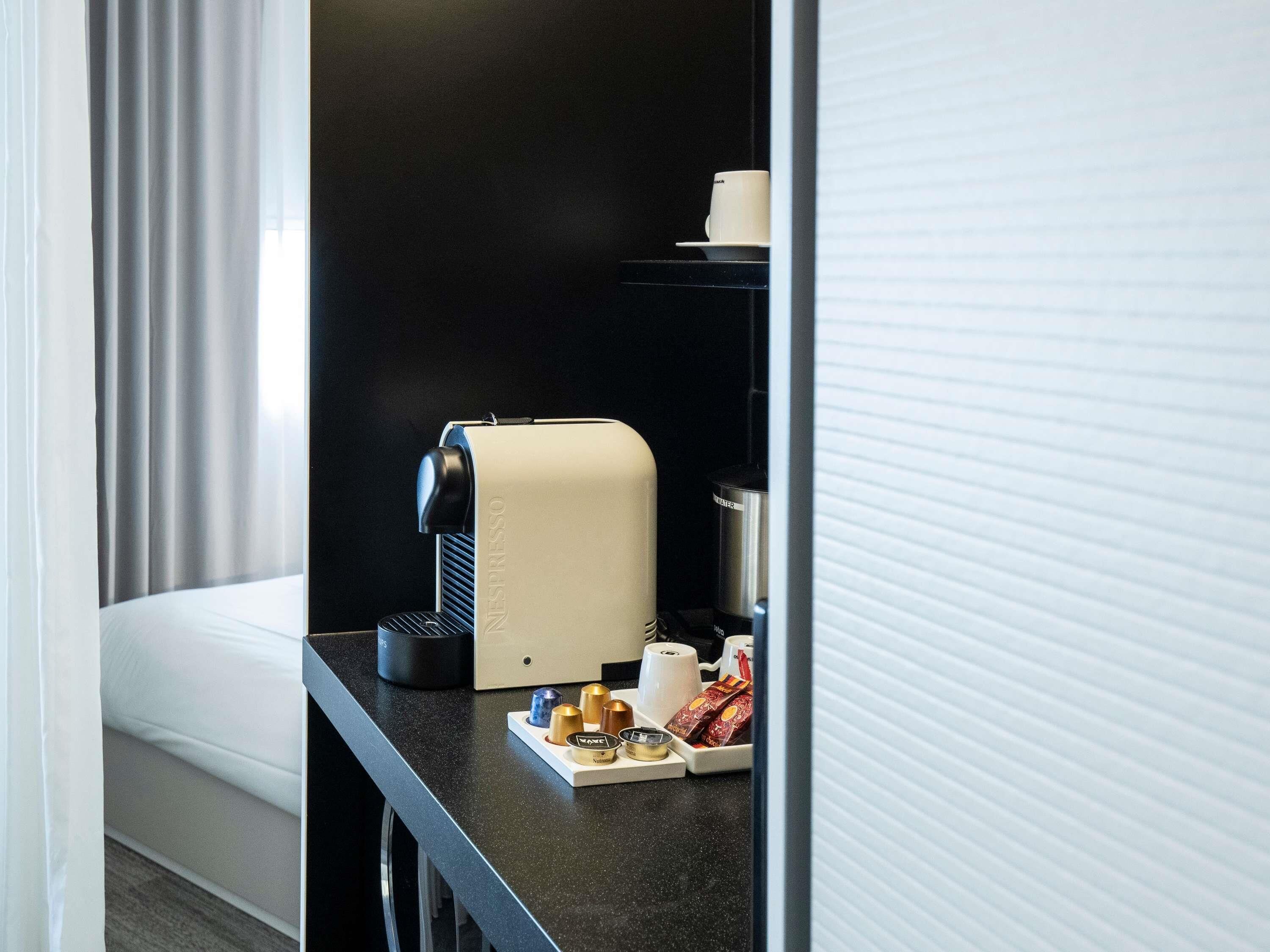 Ett hotellrum med en svart bänkskiva, en Nespresso-kaffemaskin, en kopp kaffe och ett bricka med kaffekapslar och en liten behållare.