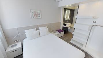 Una habitación de hotel moderna con una cama grande, una mesita de noche y un pequeño escritorio con una lámpara. Se ve un baño al fondo con un espejo y un lavabo.