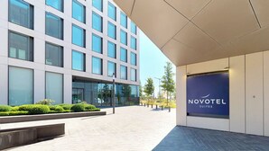 Una entrada de hotel moderna con un camino despejado hacia el edificio, un letrero de Novotel Suites y un área exterior bien cuidada con vegetación.