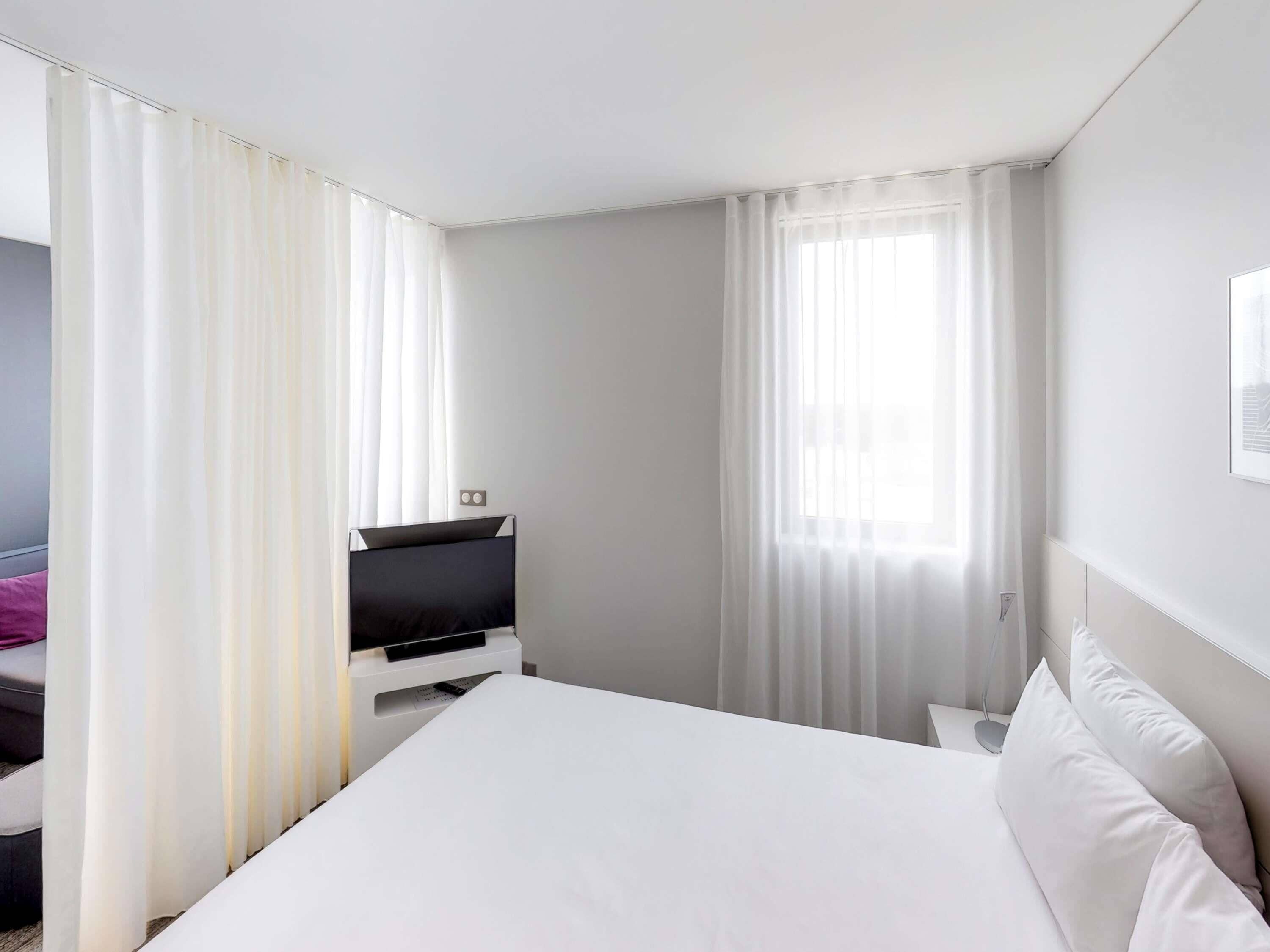 A hotel room with a bed, a TV, and a window with curtains.