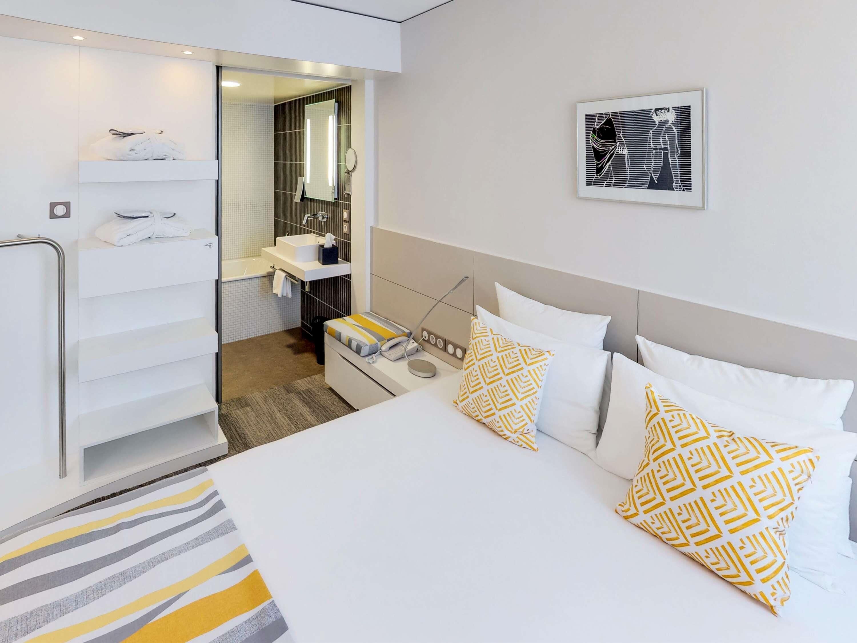 Una habitación de hotel moderna con una cama, una mesita de noche y un baño visible a través de una puerta abierta.