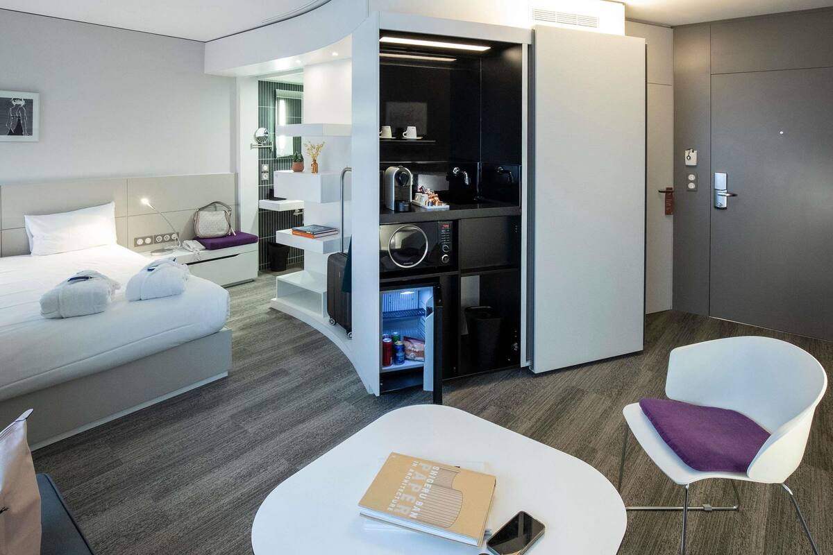 A modern hotel room with a bed, a seating area with a book and a bottle, a minibar, and a door with a lock.
