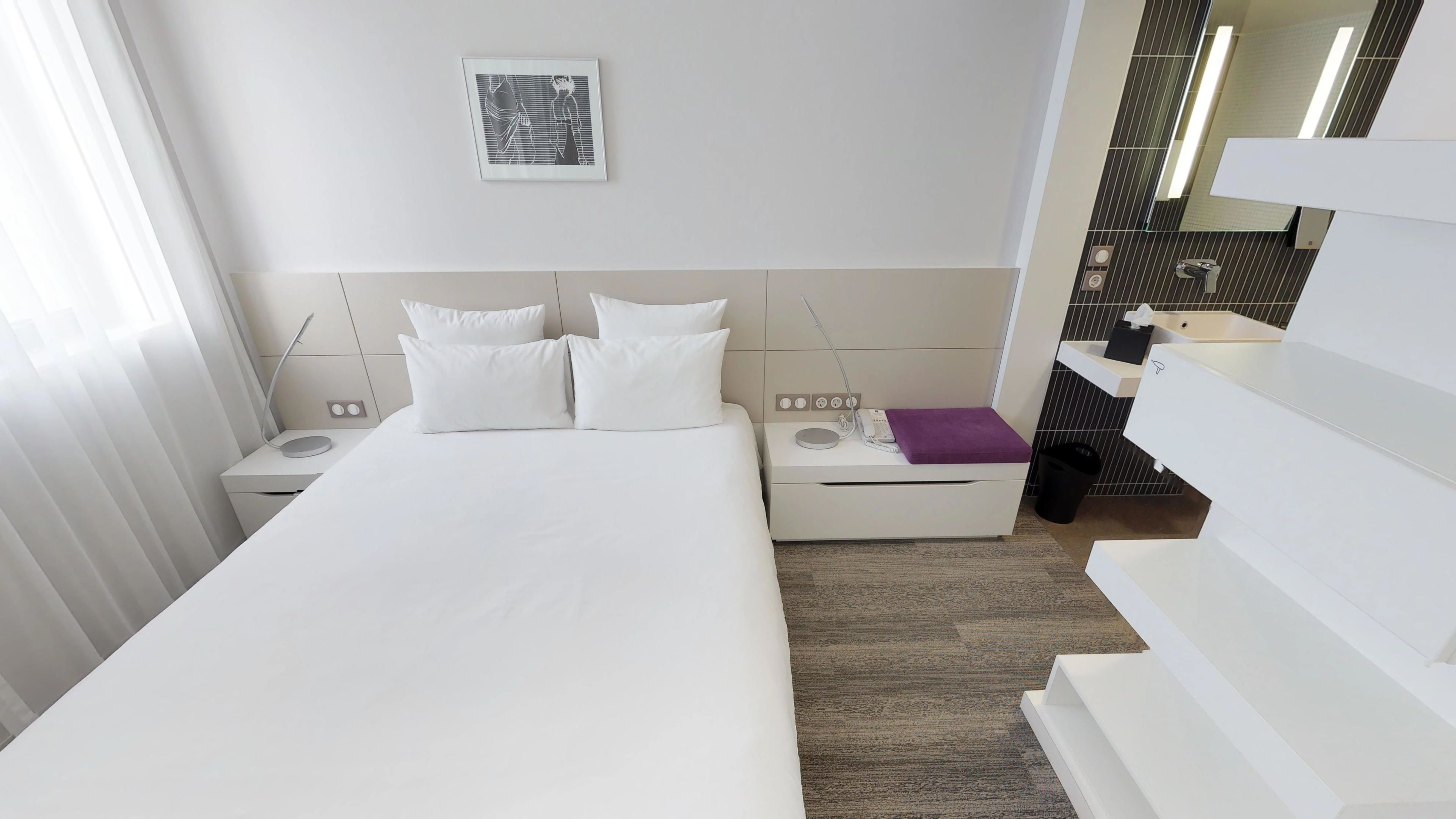 Una habitación de hotel moderna con una cama grande, una mesita de noche con una lámpara, una pequeña mesa con un teléfono y un cojín morado, y un baño con un espejo y un lavabo.