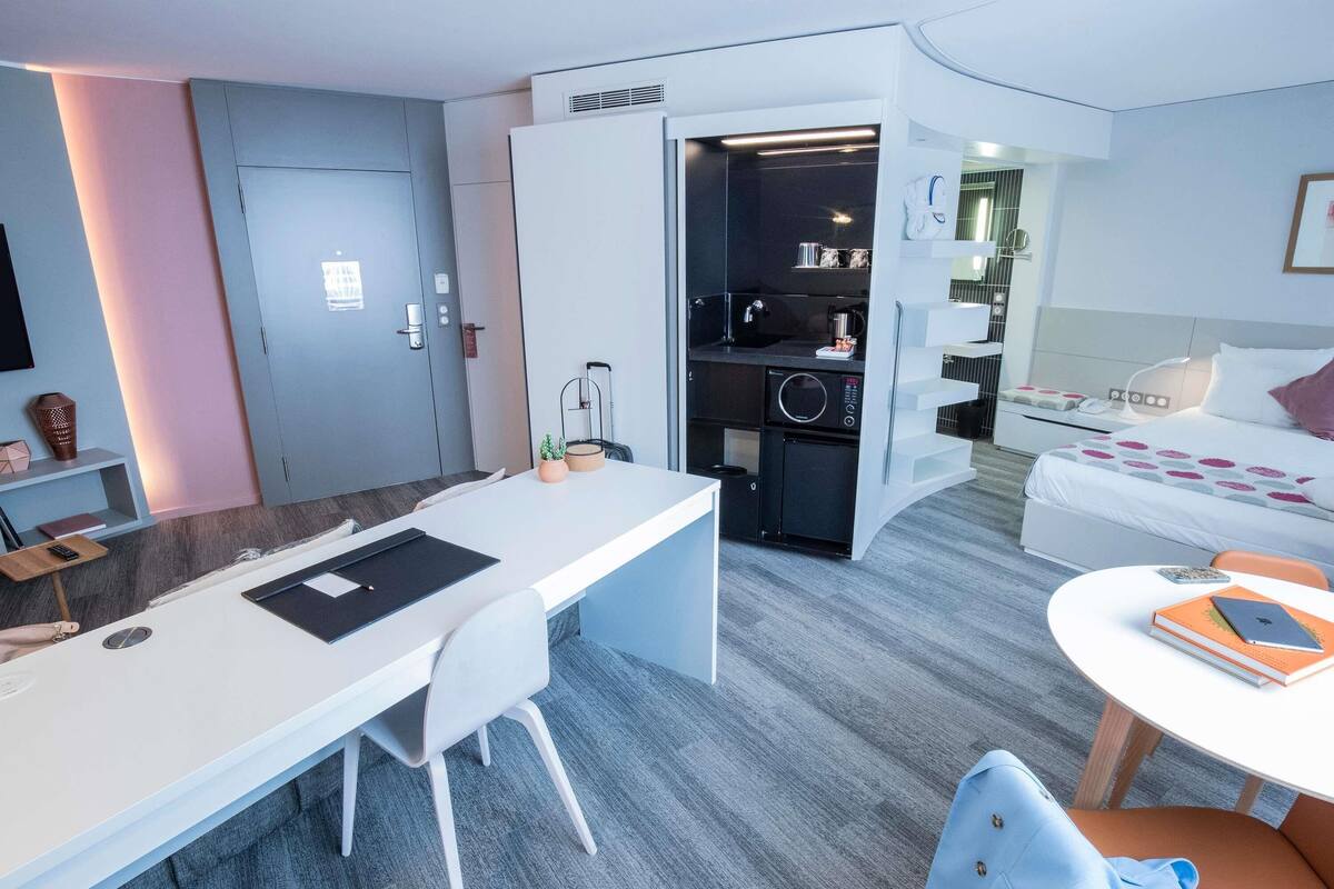 A modern hotel room with a desk, a bed, a small kitchenette, and a bathroom.