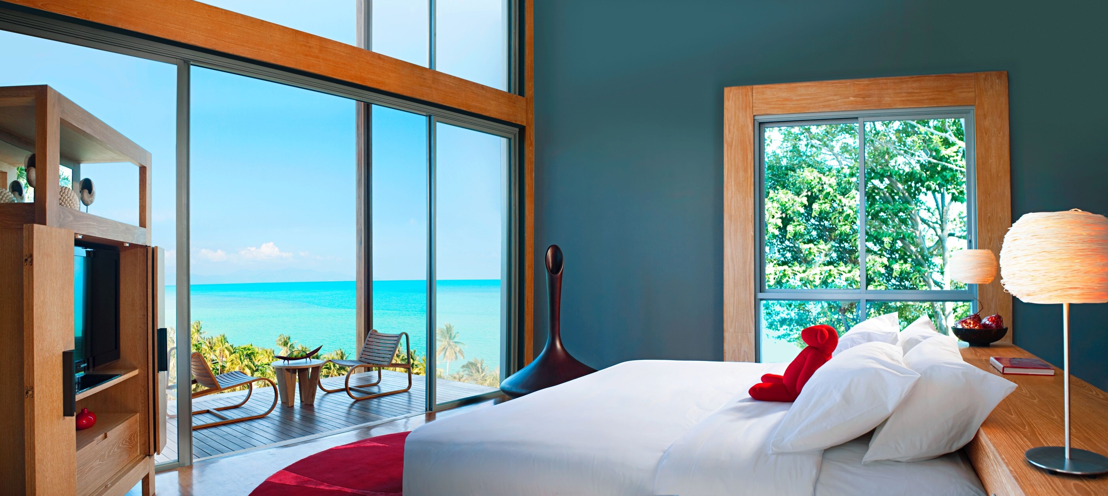 A bedroom with a large window overlooking the ocean, a bed with white linens, a red teddy bear, and a wooden nightstand.