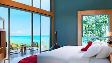 A bedroom with a large window overlooking the ocean, a bed with white linens, a red teddy bear, and a wooden nightstand.