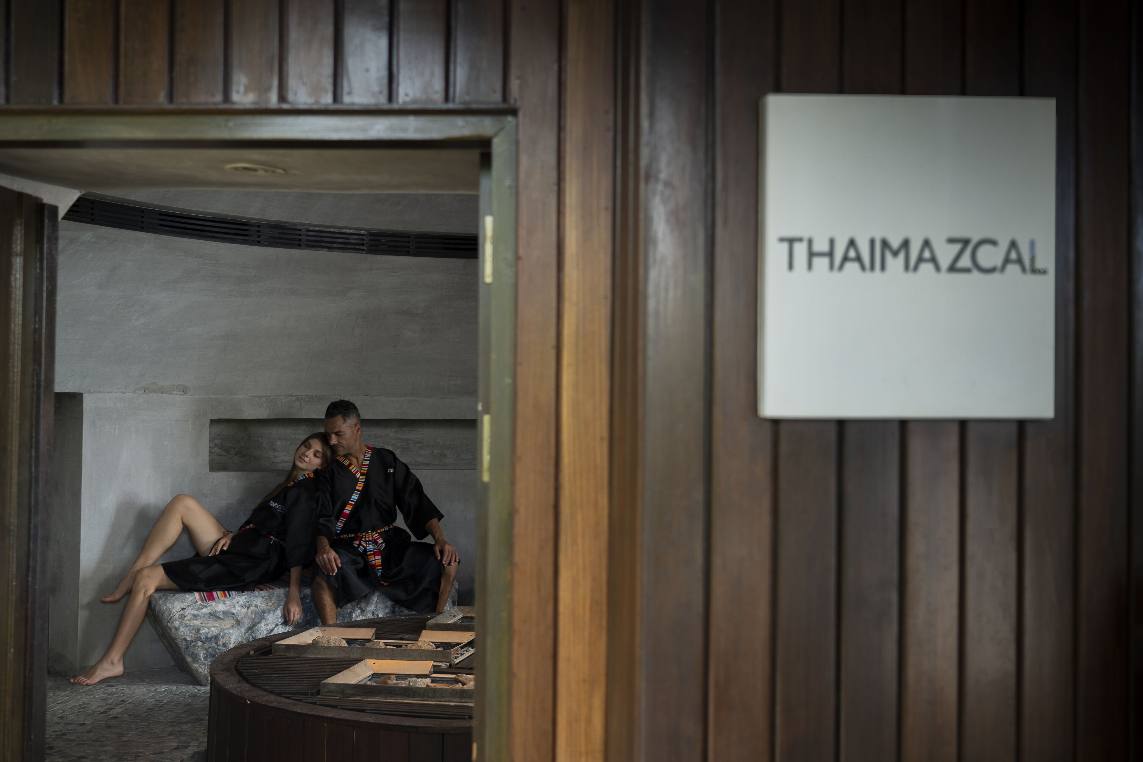 A couple relaxing on a stone bench in a spa-like setting with a sign reading "THAIMAZCAL" on the wall.