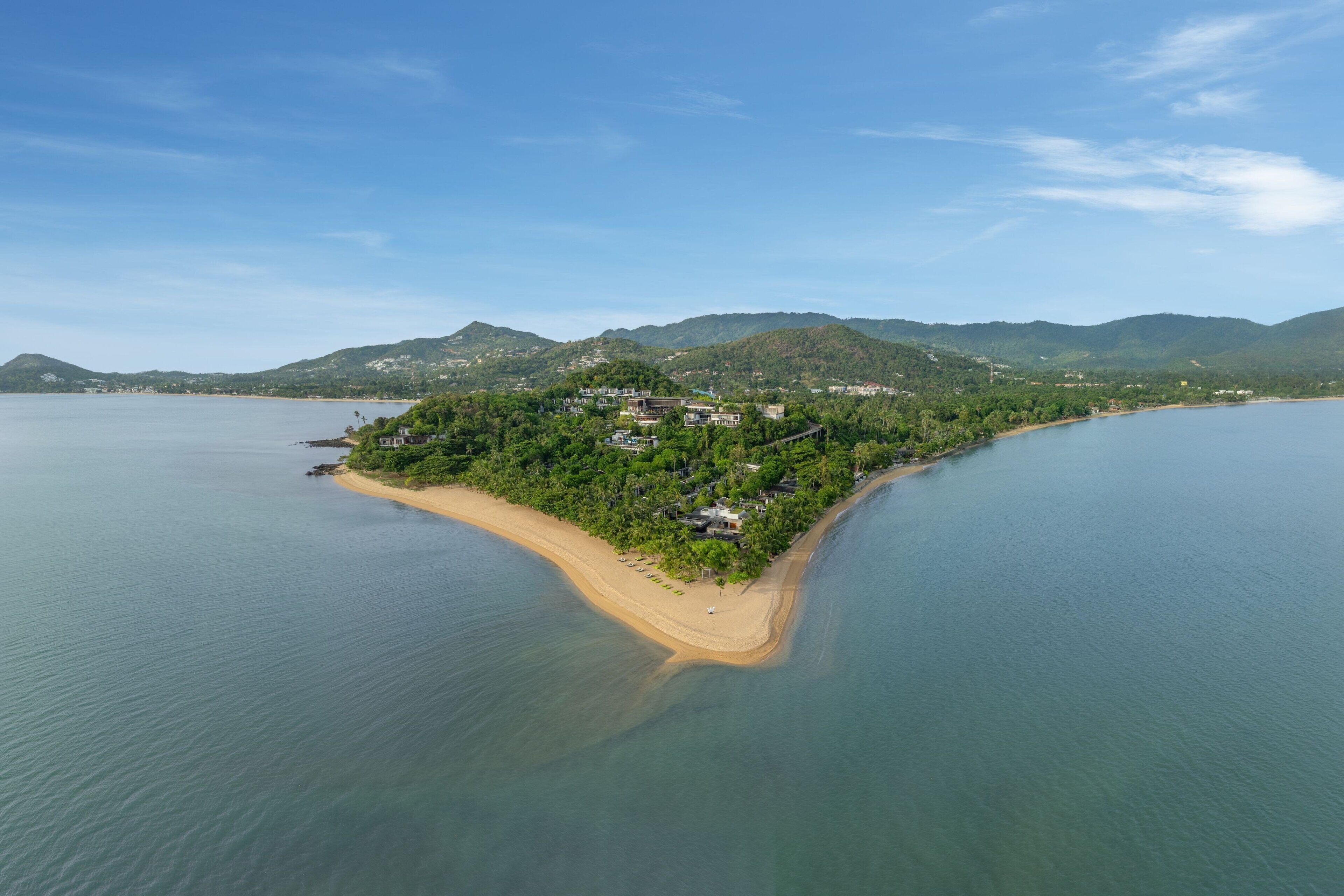 An aerial view of a coastal island with a sandy beach, lush greenery, and a few buildings.