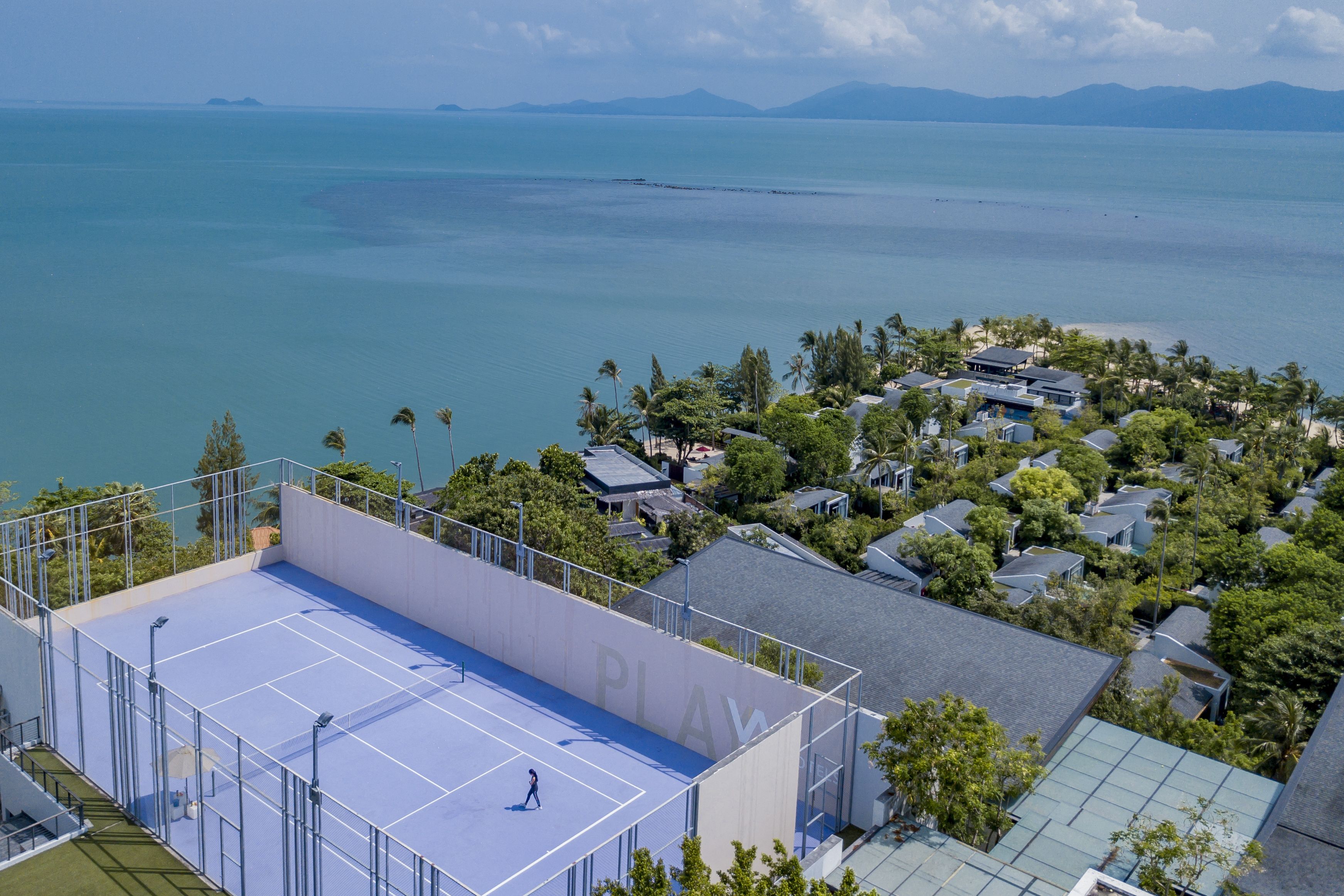 Vista aérea de una zona residencial con cancha de tenis, casas y una playa al fondo.