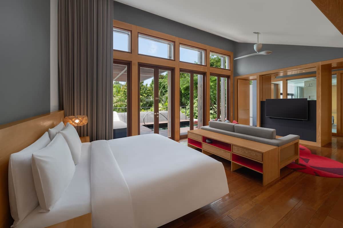 A modern hotel room with a large bed, a TV, a seating area, and large windows offering a view of the outdoors.
