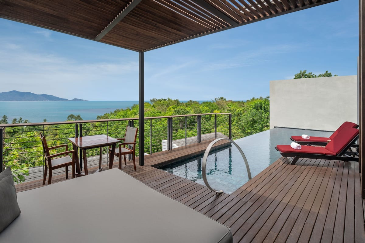 A wooden deck with a pool, red lounge chairs, and a table with chairs.