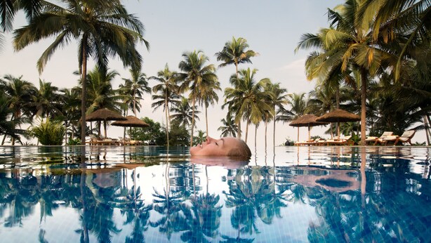 A swimming pool with clear reflections of palm trees, surrounded by lounge chairs and umbrellas. - Neeleshwar Hermitage (Hosdurg)