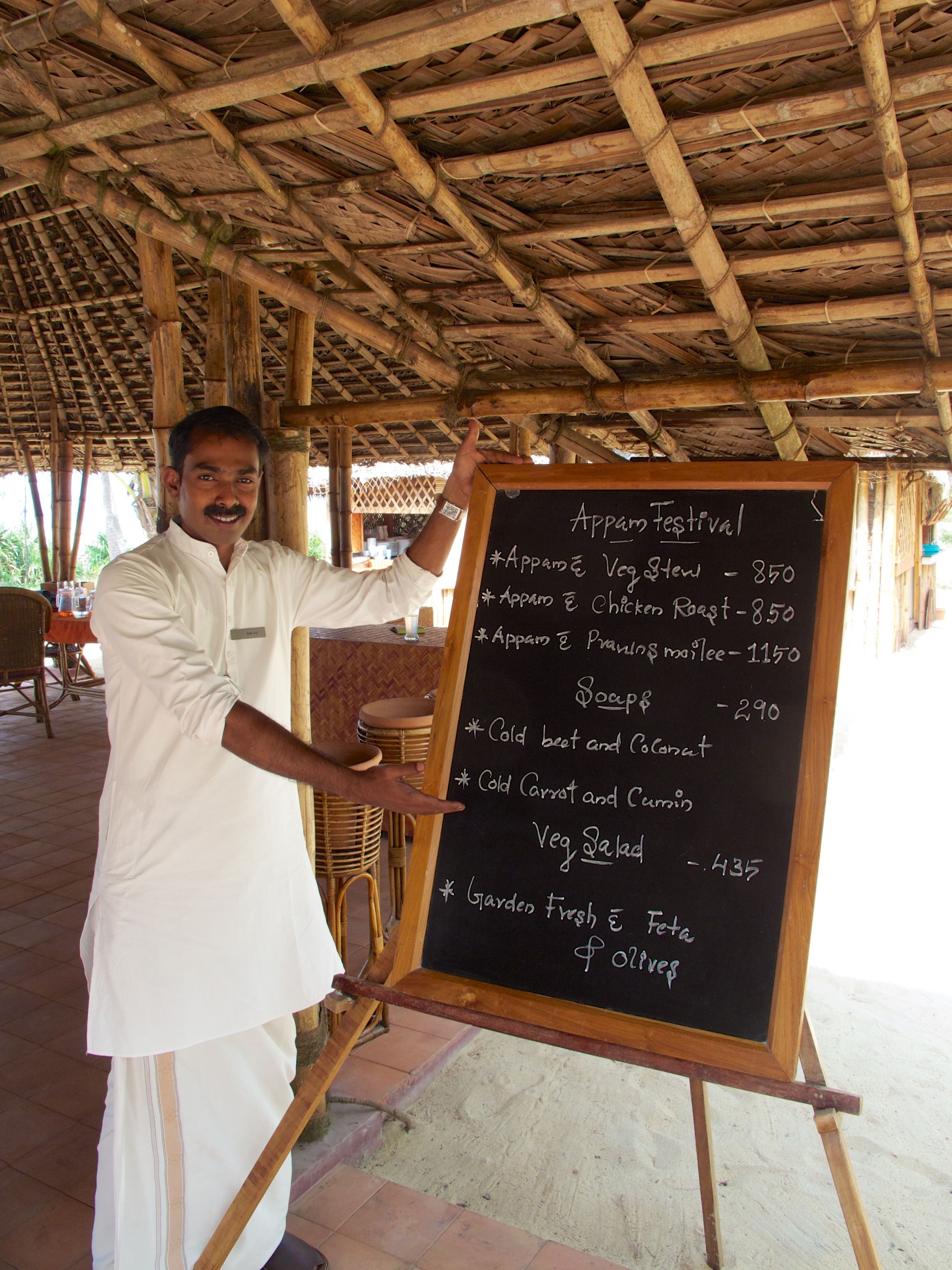 A man standing in front of a chalkboard menu listing various dishes and their prices.