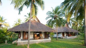 Two thatched-roof huts with wooden furniture, surrounded by palm trees and grass. - Neeleshwar Hermitage (Hosdurg)