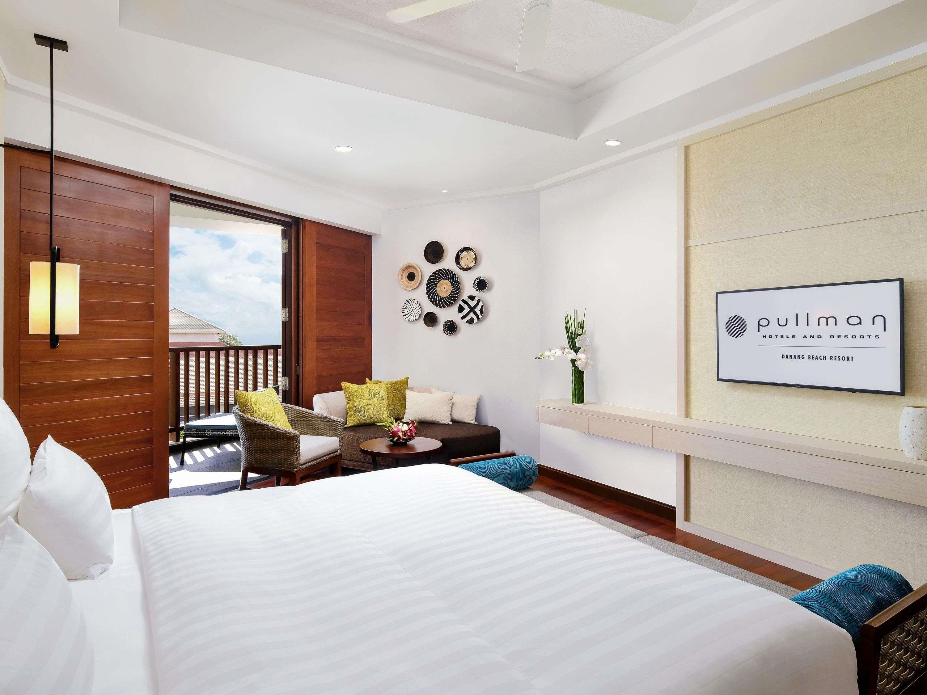 A hotel room with a bed, a seating area with chairs, a small table, a wall-mounted TV, and a balcony with a view.