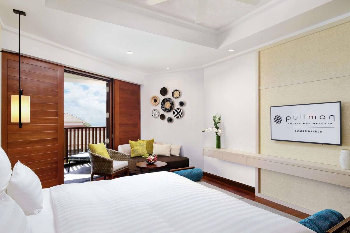 A hotel room with a bed, a seating area with chairs, a small table, a wall-mounted TV, and a balcony with a view.
