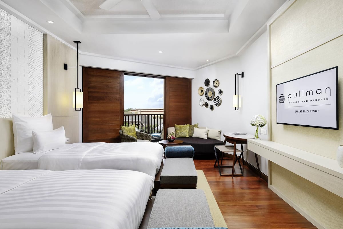 Quarto de hotel com duas camas, varanda, área de refeições e televisão fixada na parede exibindo o nome do hotel.