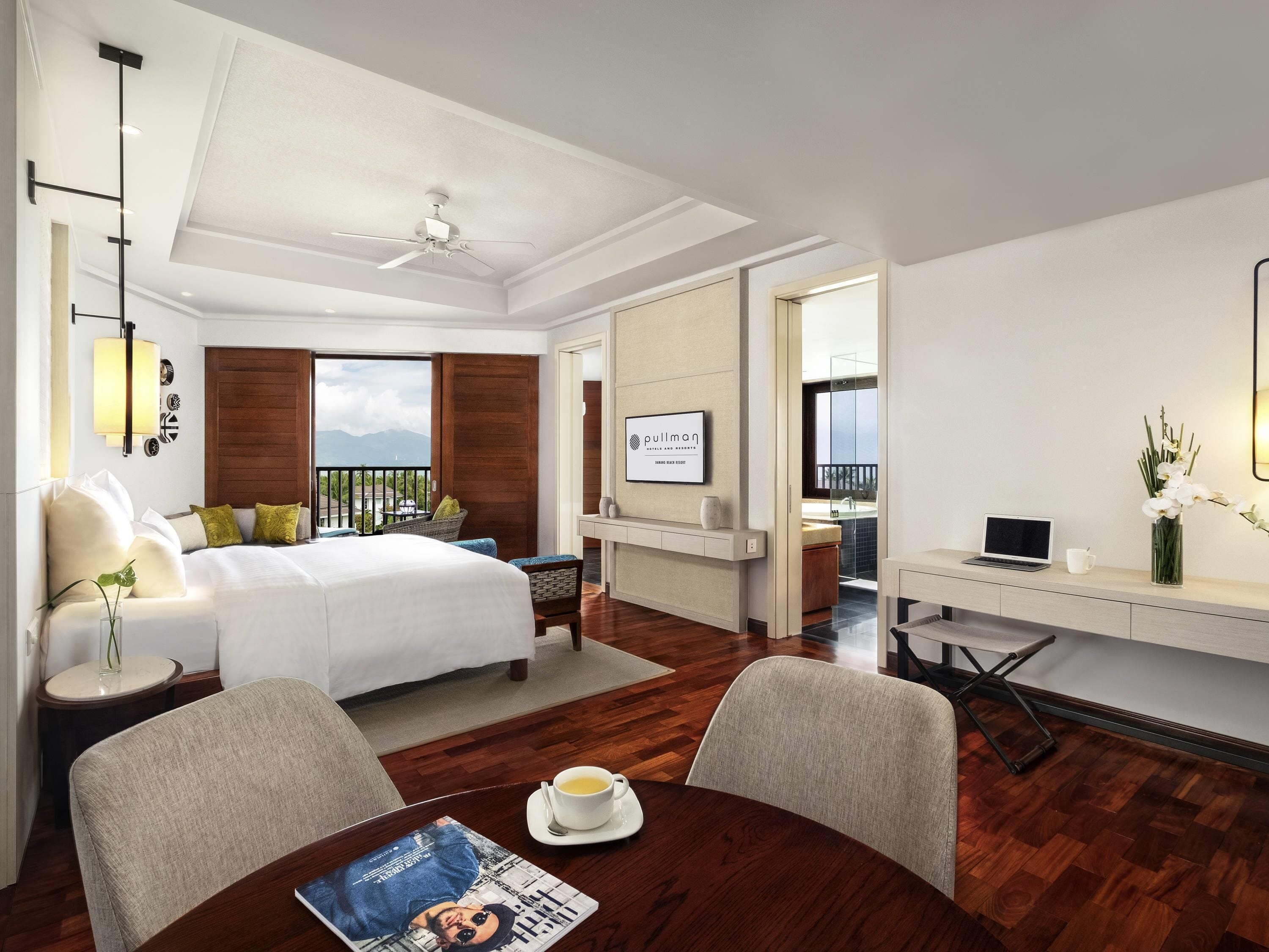 A hotel room with a bed, a desk, a chair, a table with a magazine, a cup, and a laptop.