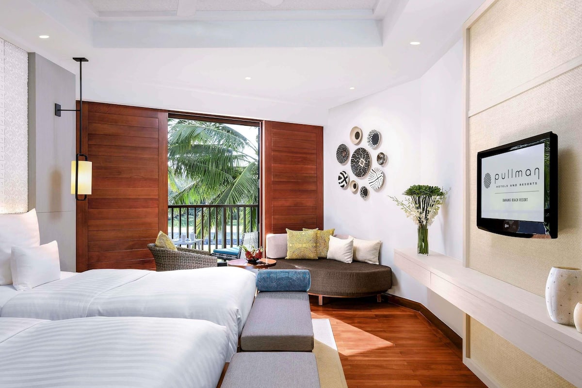 A modern hotel room with two beds, a balcony with a view of palm trees, a wall-mounted TV, and decorative wall art.