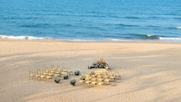A beach wedding setup with chairs arranged in a semi-circle, a floral arrangement, and a backdrop of the sea and distant mountains.