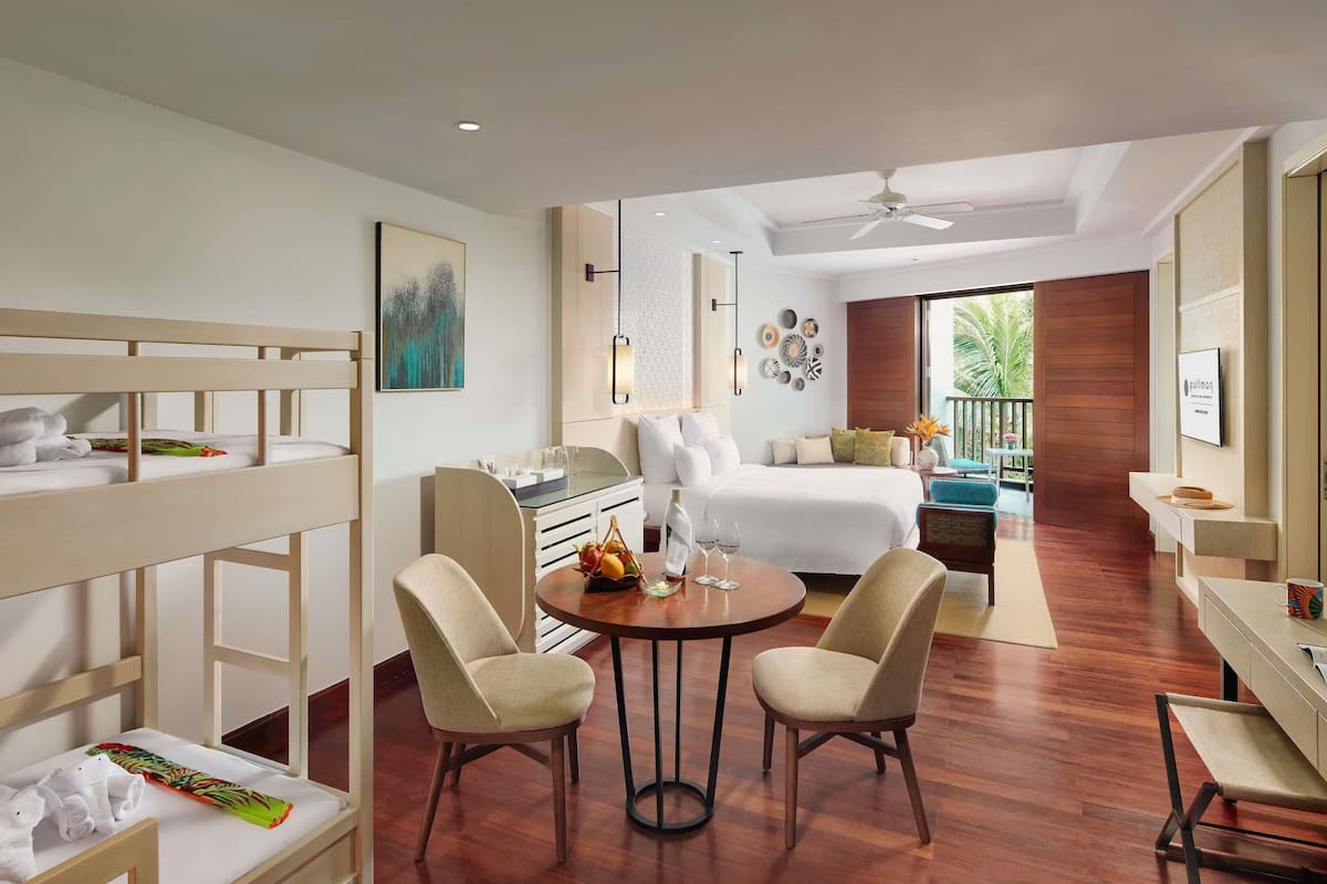 A hotel room with a bunk bed, a dining area with two chairs, a small table with a fruit basket and wine glasses, a bed with white linens, a ceiling fan, and a view of the outdoors.