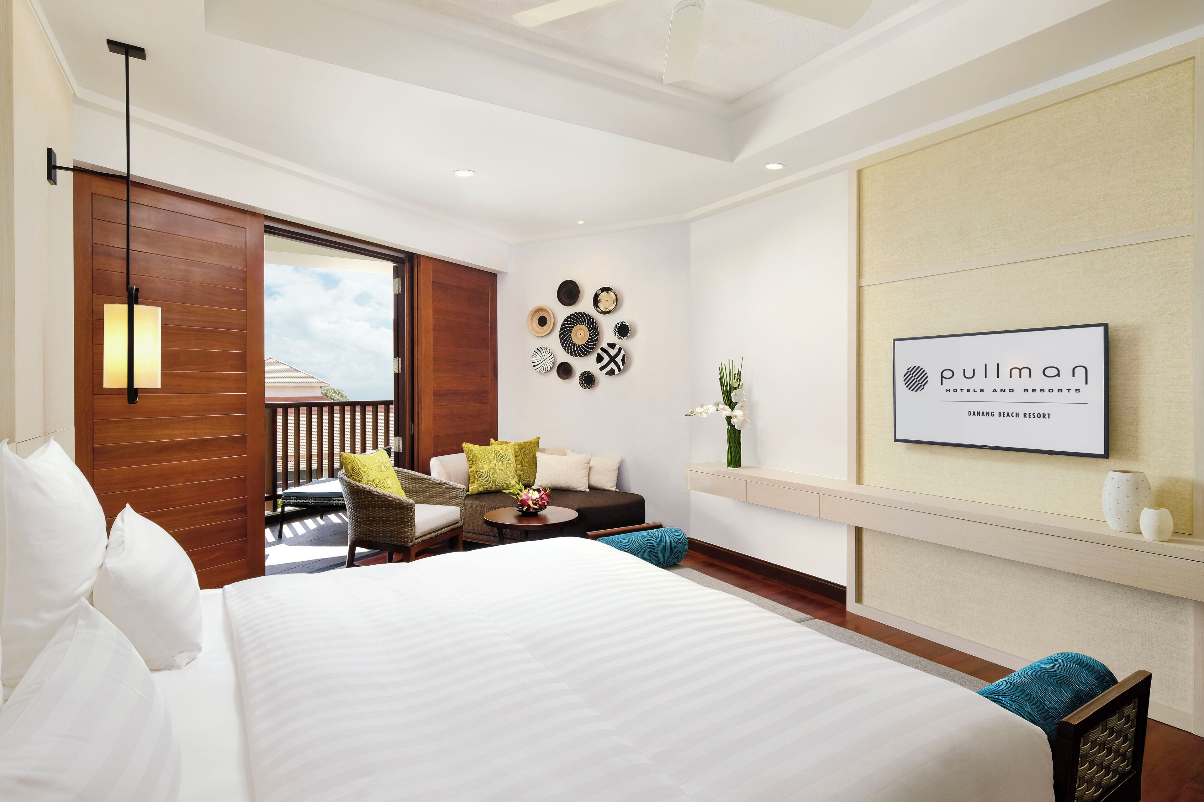 A hotel room with a bed, a seating area with a table, a wall-mounted TV, and a balcony with a view.