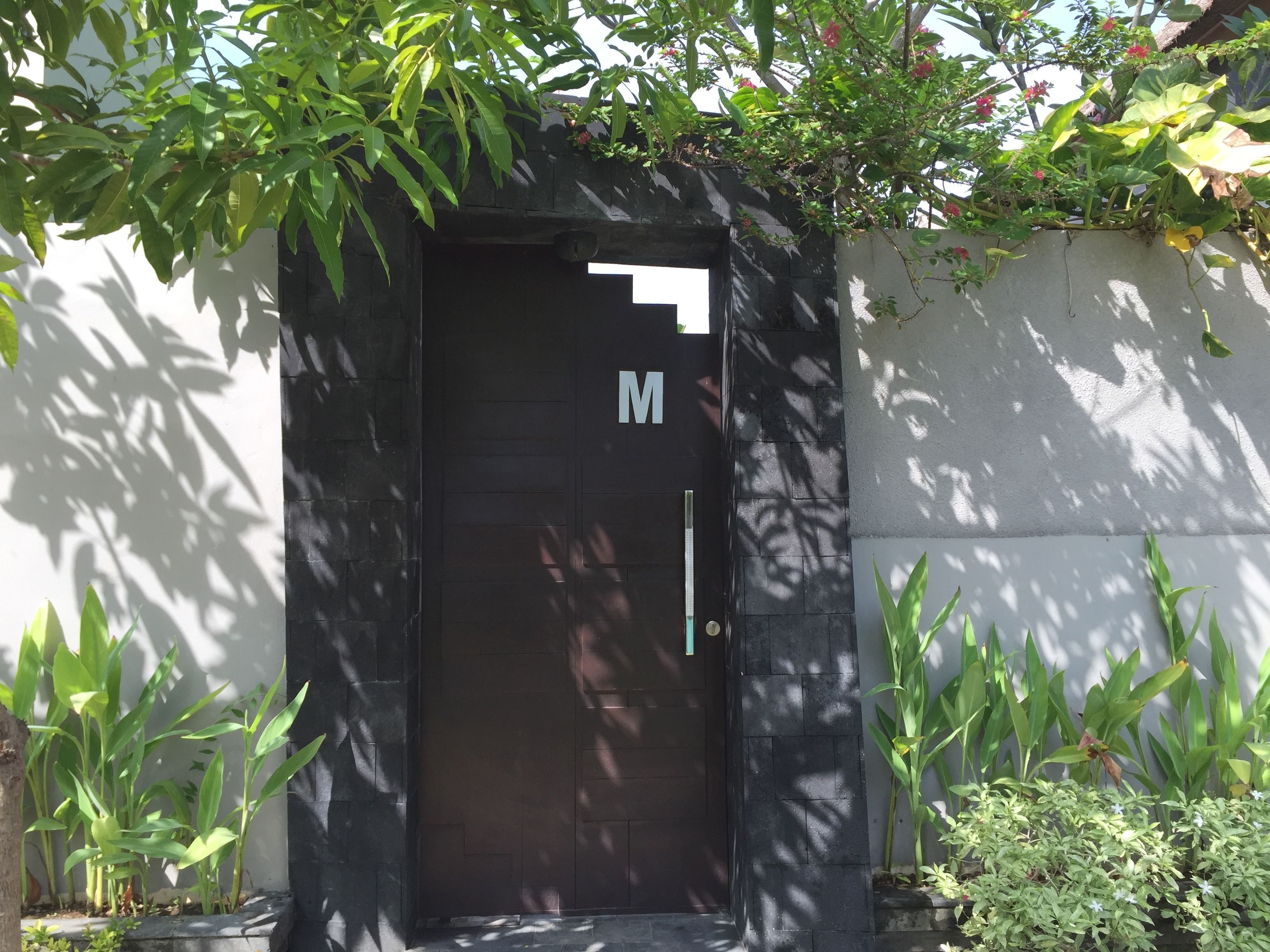 A black door with a white 'M' sign above it, surrounded by greenery and a white wall.