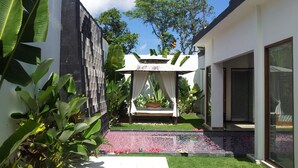 Un área de estar al aire libre con diseño moderno, con un dosel blanco, rodeada de abundante follaje y una fuente decorativa.