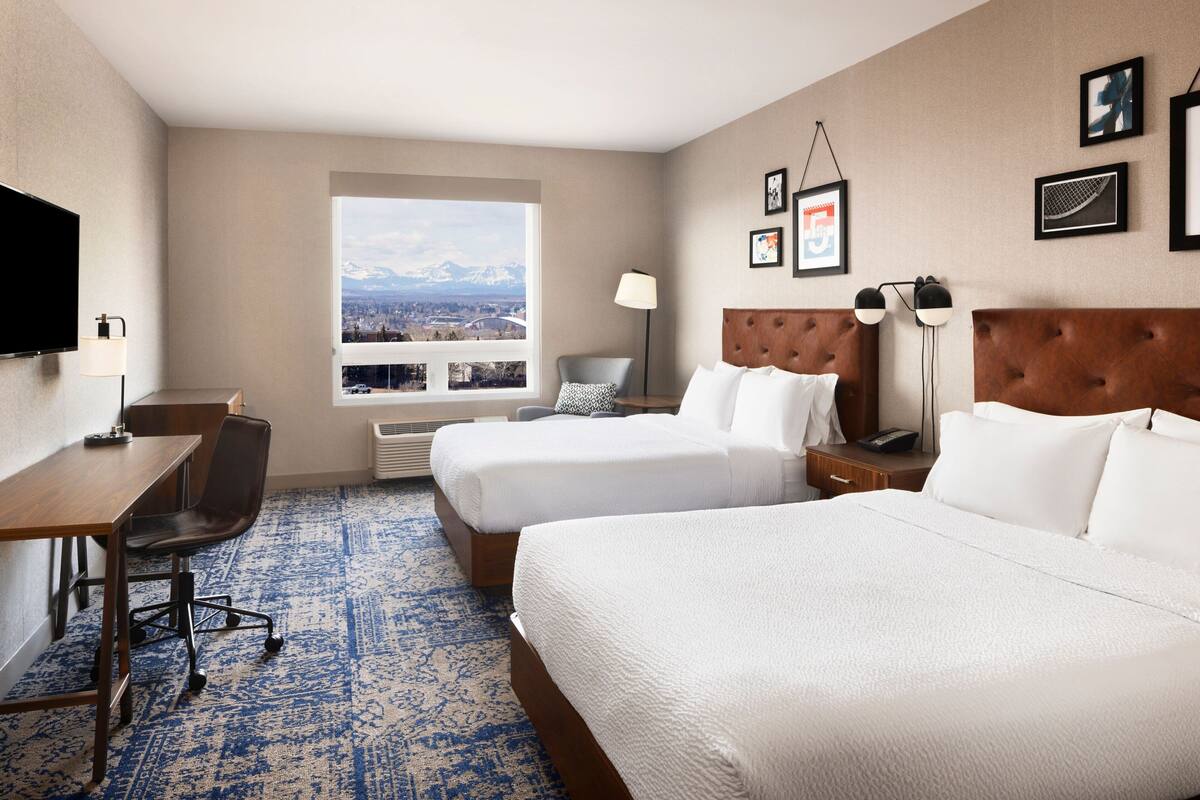 A hotel room with two beds, a desk, a chair, a TV, and a window with a mountain view.