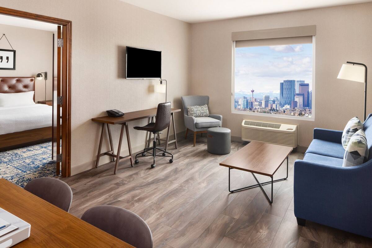 A hotel room with a bed, desk, chair, sofa, and a large window with a city view.